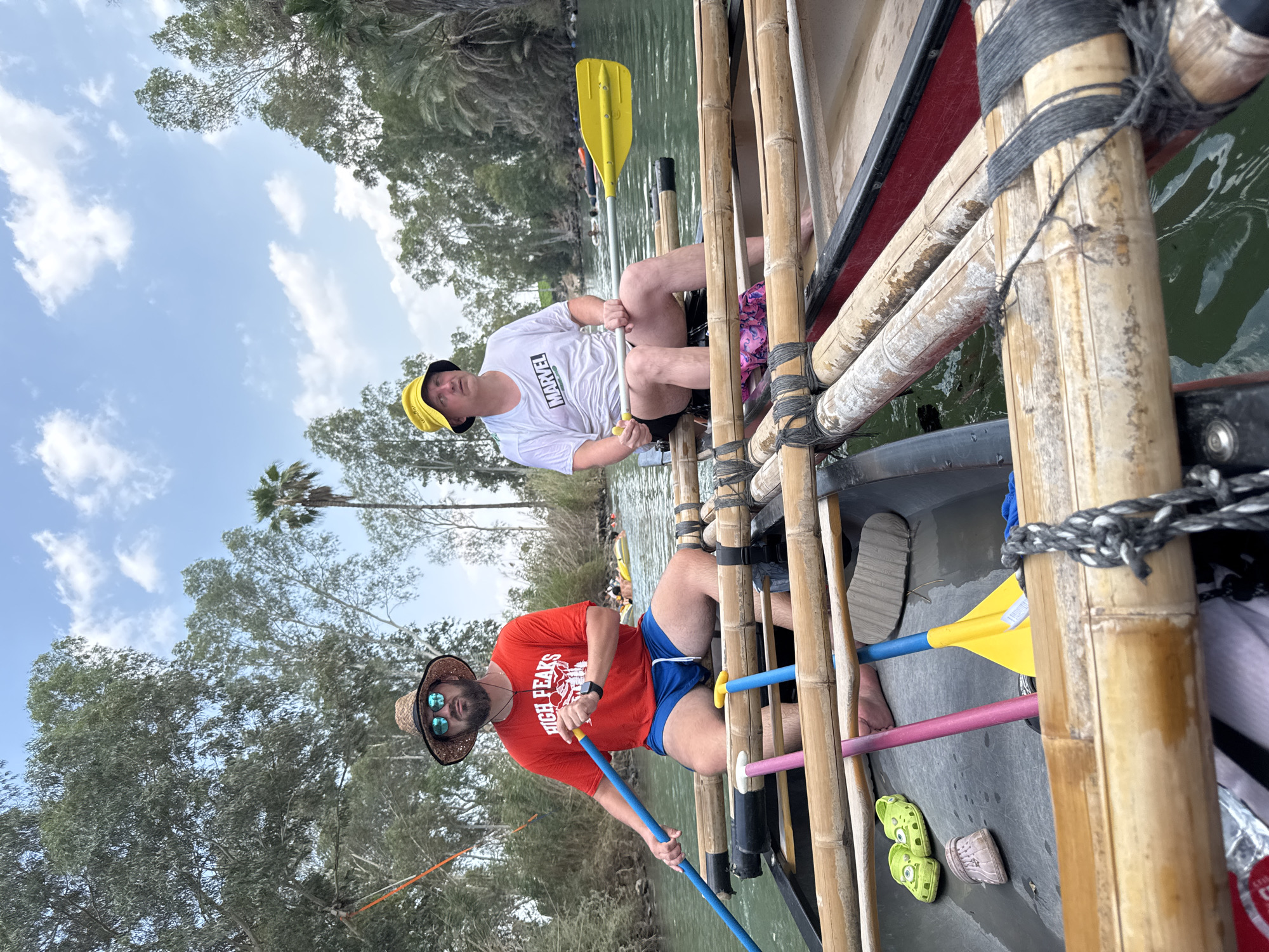 Two men are in a raft on a river in Emek HaYarden Regional Council, Israel. The man in the front is wearing a straw hat, sunglasses, a red t-shirt that says "HIGH PEAKS," and blue shorts. He is holding a blue paddle. The man behind him is wearing a yellow bucket hat, a white t-shirt with a Marvel logo, and black shorts. He is holding a yellow paddle. The raft is made of bamboo and is floating on a green river. There are trees and palm trees on the banks of the river. The sky is blue with some clouds.