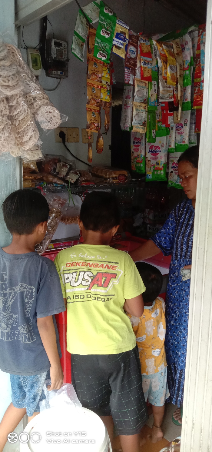 Three children and a woman are in a small convenience store. The store sells bagged snacks and treats such as "Malkist" and "Merries." One child wears a blue "Work Zone" t-shirt, while another wears a lime green t-shirt. The woman is wearing a blue patterned dress. A bag of chips hangs from the store's ceiling. The store has an electric meter and is stocked with various food items.