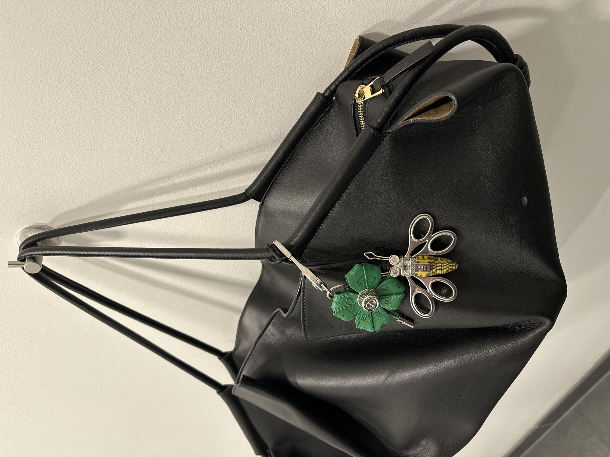 A black leather handbag hangs from a brushed metal hook on an off-white, textured wall in the City of London, United Kingdom. The bag, featuring two narrow black leather straps, exhibits soft folds and an open section revealing a gold-toned zipper. Two distinct keychains are attached via a silver-toned clasp to one of the straps. The first is a green, flower-shaped retractable tape measure with numerical markings and a central "CD" logo. The second is a silver-toned bee-shaped charm, its body striped yellow and black, resembling a miniature tape measure, with silver loop wings. A dark horizontal line is visible at the bottom of the frame, indicating a floor or base. No people are present.