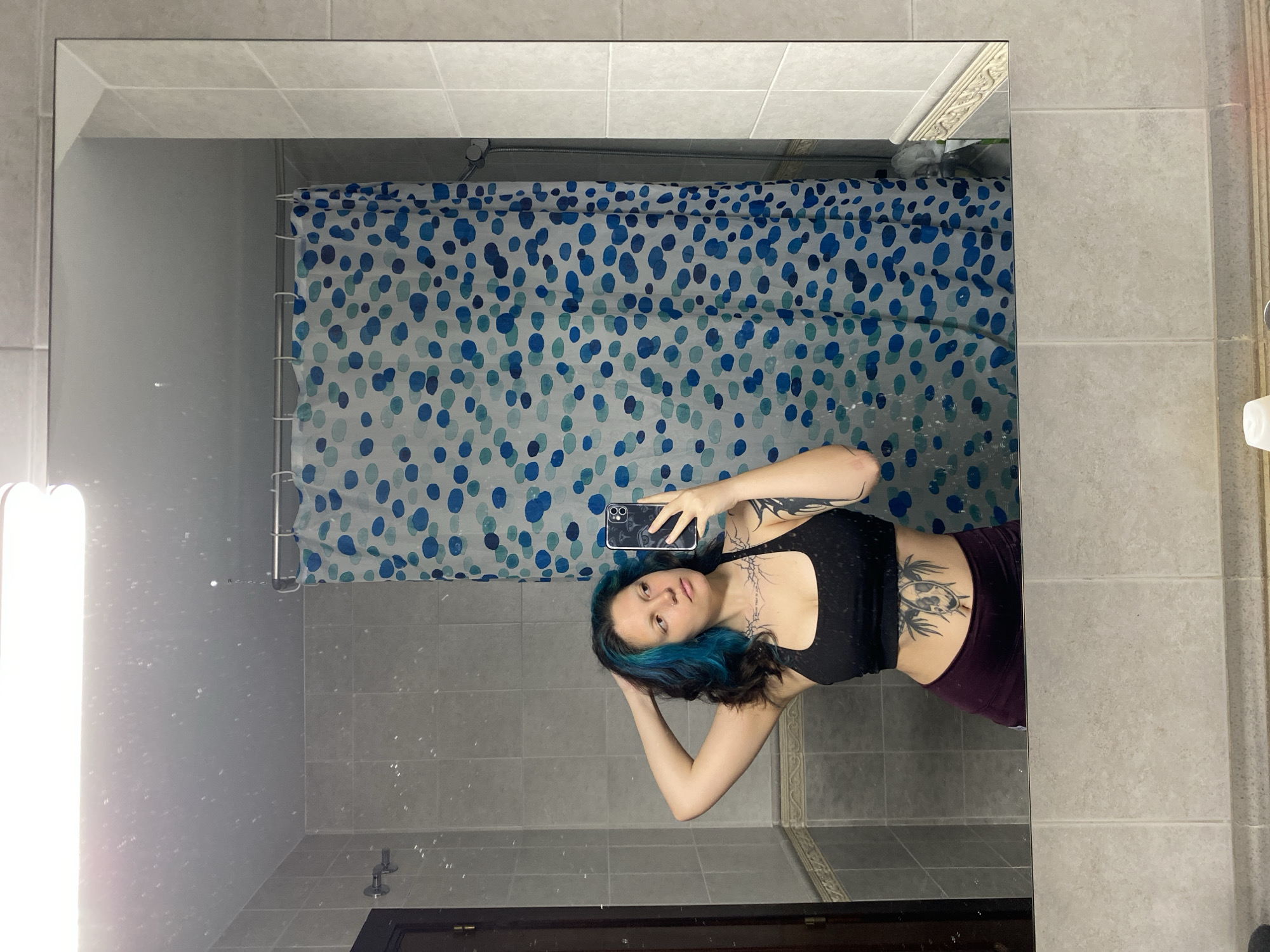 A person with blue-tinted hair and several tattoos is taking a selfie in a bathroom mirror. The bathroom has a tiled wall and a shower curtain with blue and white circular patterns. The person is wearing a black tank top and purple pants. A smartphone is visible in the person's hand. The bathroom's door, along with the bathroom amenities, are observed within the mirror reflection. The location is unknown but the prompt mentions the city of Gijón/Xixón, Spain.