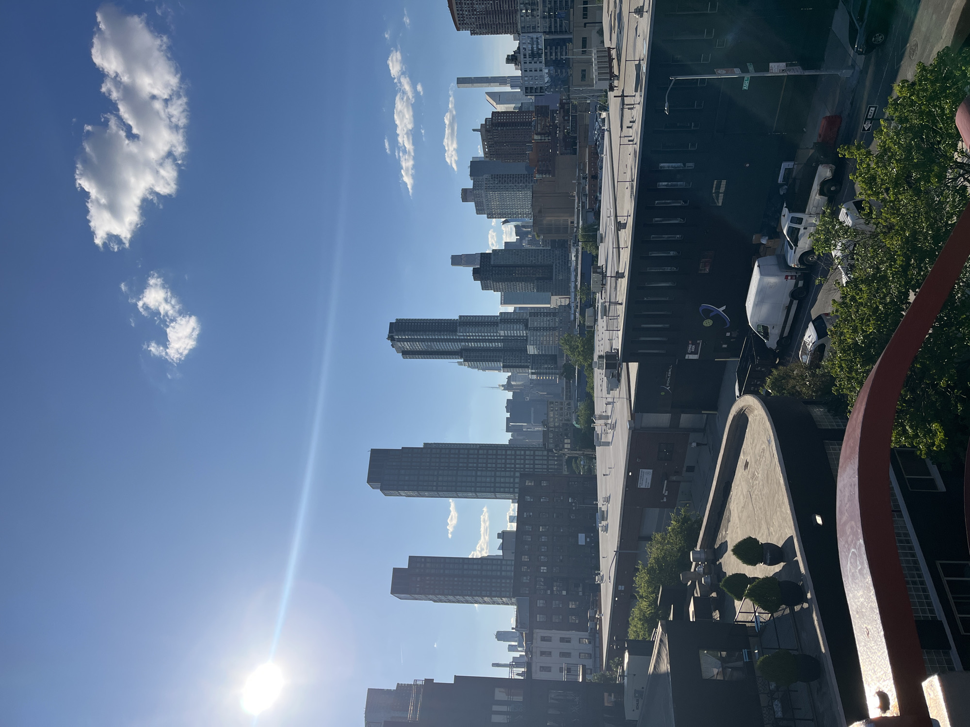 The image displays a cityscape of New York City, likely from an elevated vantage point, possibly a rooftop. The foreground features the top portion of a reddish-brown structure, possibly a railing or architectural element, partially obscuring the view below. Beyond this, several flat-roofed buildings are visible, one with a curved edge, and topped with potted plants. In the mid-ground, a cluster of industrial-style buildings with numerous windows and skylights dominate the scene. Further in the background, a dense collection of skyscrapers rises, showcasing a variety of architectural styles and heights, indicative of a major urban center. The sky is a clear blue with a few scattered white clouds, and the sun is bright, casting lens flare and illuminating the scene. The presence of distinct skyscrapers and the overall urban density strongly suggests this is a view within one of New York City's boroughs, possibly with Manhattan's skyline in the distance.
