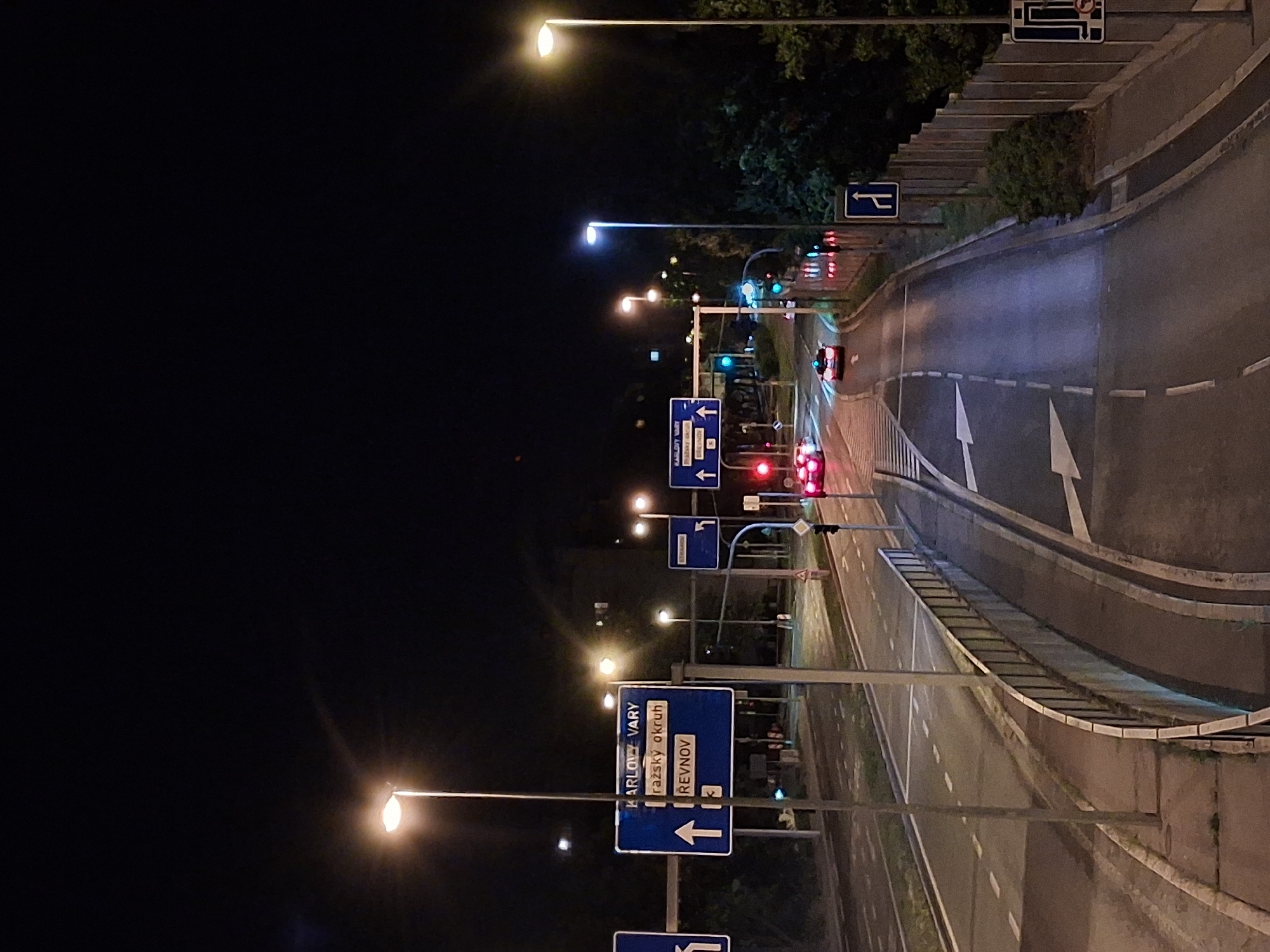 The image depicts a street scene at night in Prague, Czechia. The perspective is from a slightly elevated viewpoint looking down at a multi-lane road. There are several directional signs with white text on blue backgrounds indicating destinations such as "KARLOVY VARY," "pražský okruh," and "ŘEČNOV." Streetlights illuminate the roadway and surrounding area. Cars are visible on the road, some with their headlights on, and traffic signals are present. On the right side of the image, a tall fence runs along the edge of the road, with trees and foliage visible behind it. The overall environment suggests an urban setting with active traffic flow.