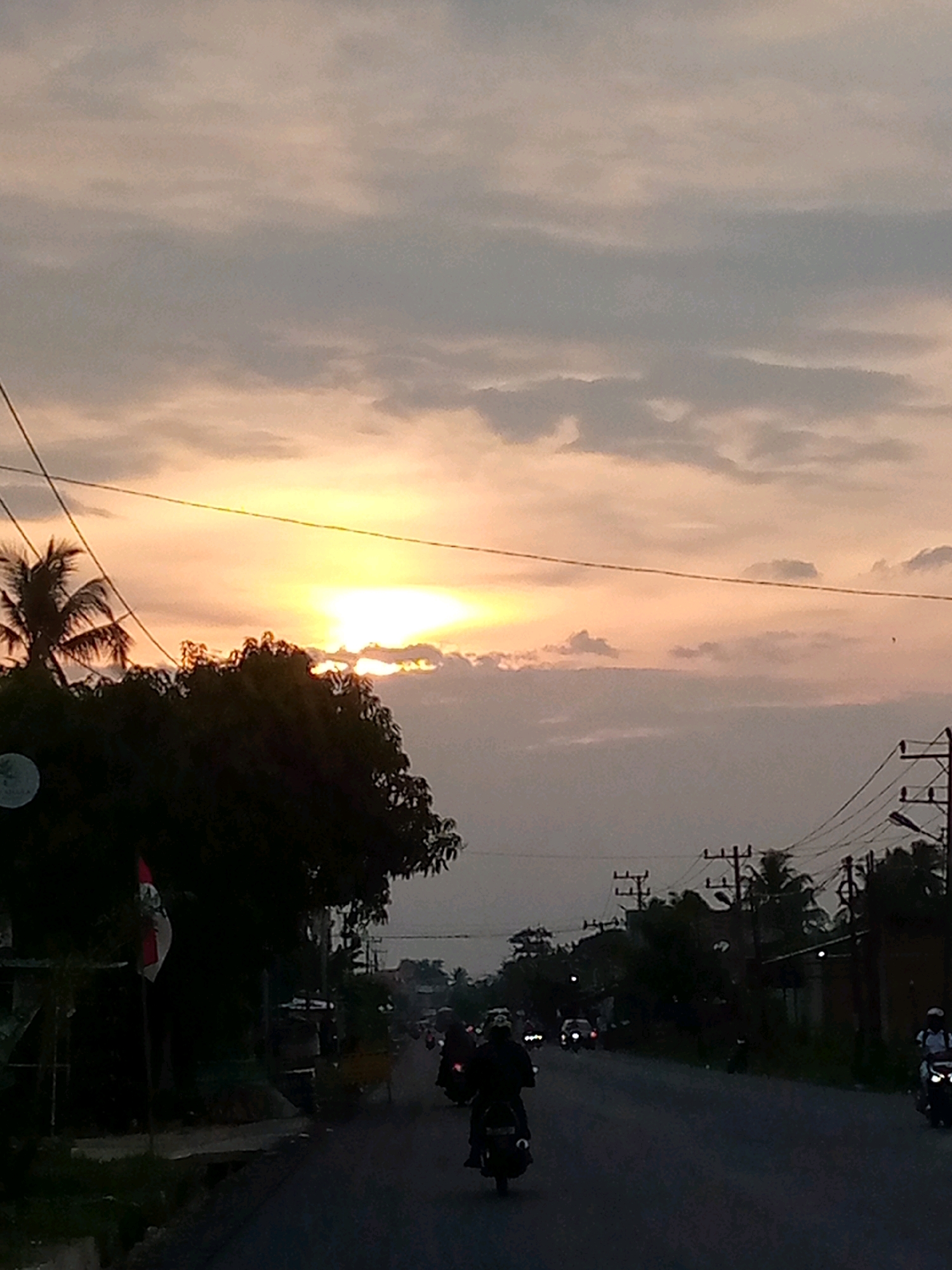 The image depicts a street scene in Keudeteupinpunti, Indonesia, during a sunset. The sky is filled with soft, diffused light from the setting sun, casting a warm glow and creating a serene atmosphere. Wispy clouds are scattered across the sky, adding texture and depth to the scene. Silhouetted trees and palm fronds are visible against the bright sky, hinting at the tropical environment.
On the street, several individuals are riding motorcycles. Their forms are partially obscured by the dimming light, but it's clear they are navigating the road. One motorcycle is particularly prominent in the foreground, with a rider wearing a dark jacket. Further down the road, more motorcycles and a few cars can be seen, their headlights starting to illuminate the asphalt.
Utility poles with wires crisscross the frame, adding a touch of urban infrastructure to the natural beauty of the sunset. A flag, possibly the Indonesian flag, is visible on a pole near the left side of the image. The overall impression is one of a typical evening commute in a coastal or rural Indonesian town, bathed in the gentle light of a setting sun.