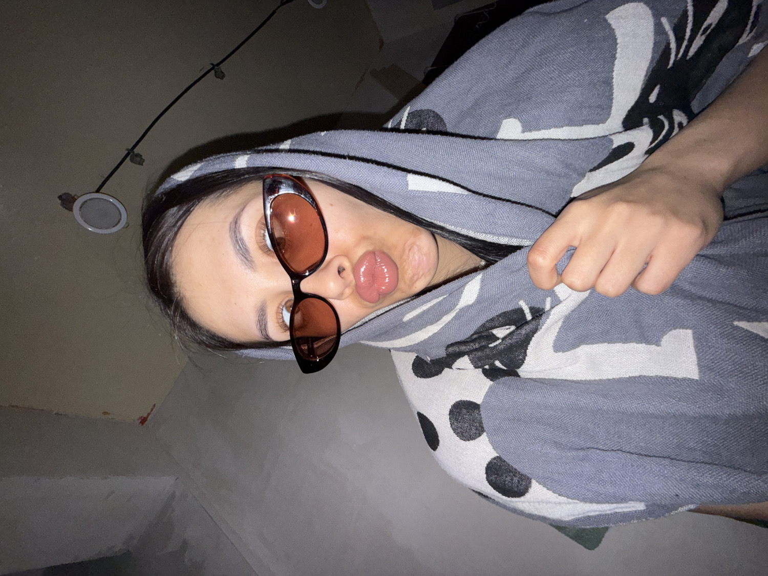 A young person is featured in a close-up, angled indoor shot, taken in Votkinsk, Russia. The subject, likely female, has dark hair and is wearing fashionable sunglasses with brown-tinted lenses and dark frames. She is making a "duck face" or pouting her lips towards the camera. She is wrapped in a soft, patterned blanket or shawl that is predominantly grey, with sections of white and black abstract shapes and polka dots. Her right hand is visible, resting on the fabric of the blanket. The background is a plain, light-colored wall or ceiling, with a single recessed circular light fixture visible in the upper left of the frame. The image's tilted orientation suggests a casual selfie or a photo taken from an unusual angle. No text is visible in the image. The overall impression is one of casualness and playfulness.