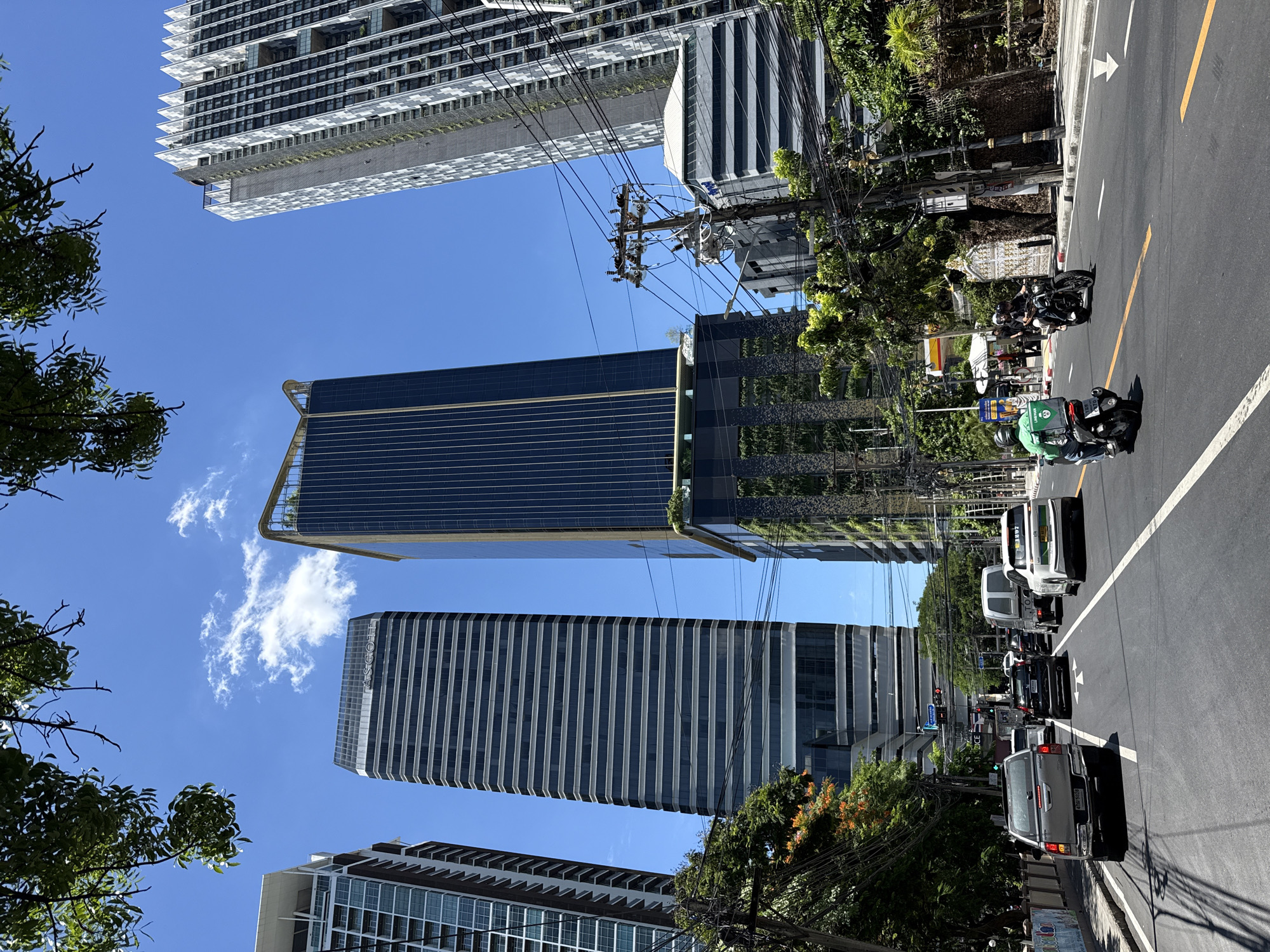 The image shows a street scene in Bangkok, Thailand. It is a sunny day with a clear blue sky and some scattered clouds. The street is lined with tall, modern buildings, including skyscrapers with glass facades. There is traffic on the road, with cars, trucks, and motorcycles. Several people are visible, including motorcyclists and pedestrians. Trees and foliage are present along the sides of the road, adding some greenery to the urban environment. The image appears to be taken from a moving vehicle, given the perspective and motion blur on some elements. The overall impression is that of a bustling city street in a tropical climate.