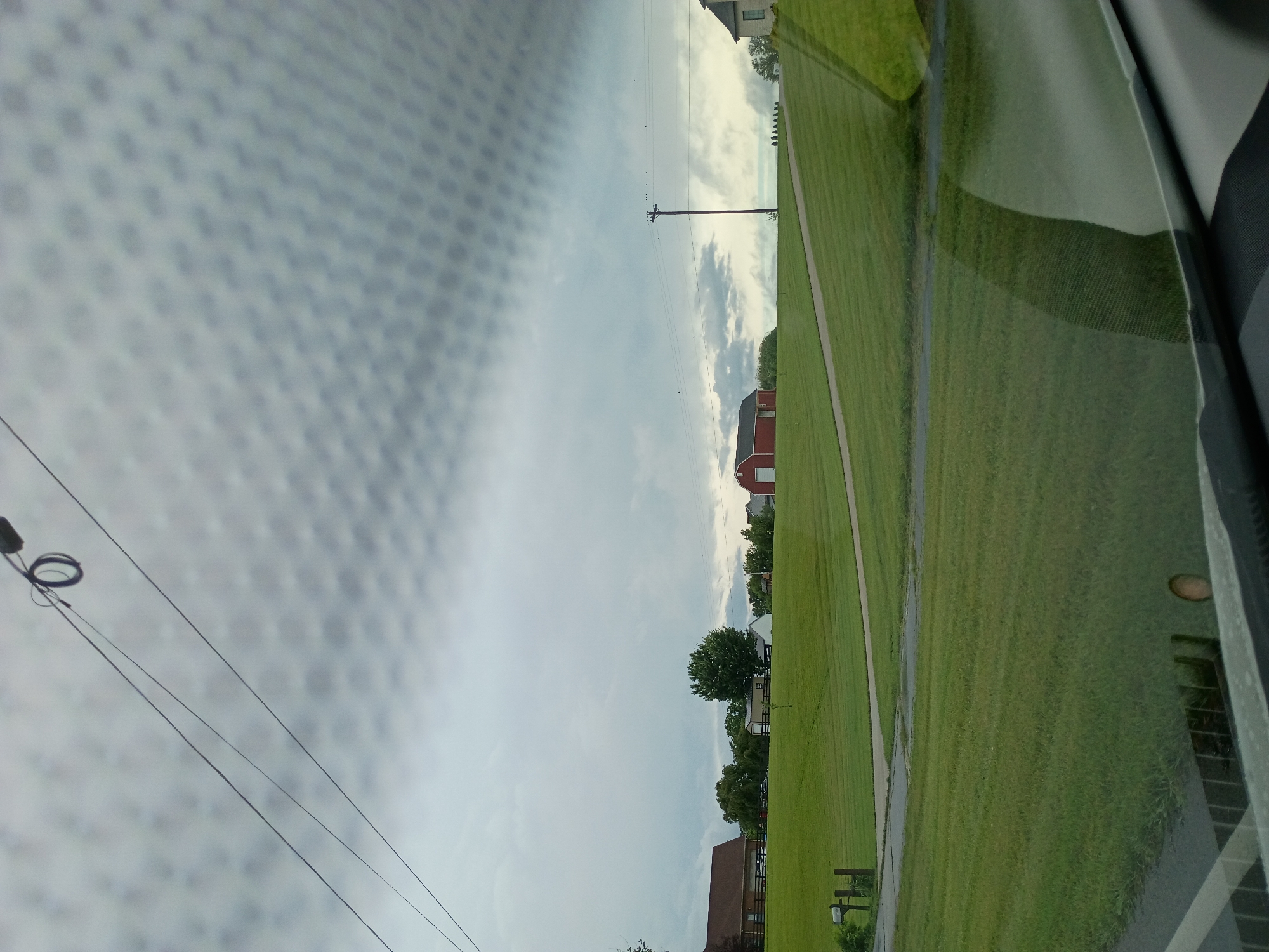 The image depicts a cloudy sky over a rural landscape, viewed through a textured surface, possibly a window with a screen. The scene is dominated by a vast expanse of green, well-maintained grass, suggesting a park, golf course, or residential lawn. A paved pathway or sidewalk runs diagonally through the grass.
In the background, a bright red barn stands out against the overcast sky, accompanied by a few other buildings that appear to be houses. A utility pole with power lines stretches across the sky, adding to the rural atmosphere. Trees are scattered throughout the scene, some appearing well-established and others more young.
The lighting suggests it's daytime, but the heavy cloud cover implies a potentially overcast or approaching storm. The texture in the foreground, possibly a window screen or a patterned surface, distorts the view of the landscape, creating a dappled effect. There is no visible text in the image. The overall mood is serene and quiet, characteristic of a rural setting.