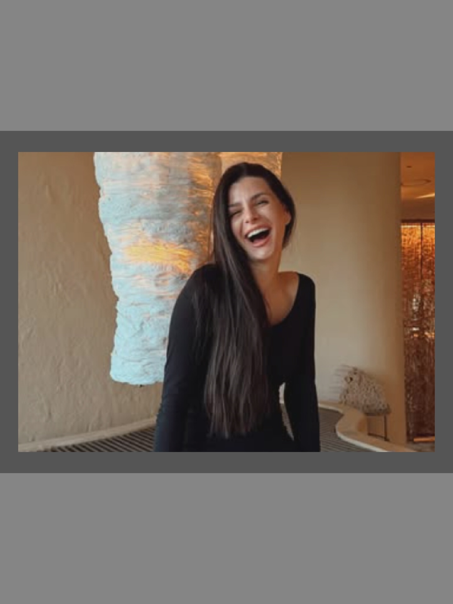 A medium shot captures a young woman with long, dark hair, wearing a black long-sleeved shirt, laughing heartily. She is positioned in the foreground against a neutral-toned wall. To her left is a large, textured, white lamp or sculpture with a warm light emanating from within. To her right, a portion of a patterned wall and a decorative object are visible. The scene appears to be indoors, possibly a restaurant or lounge, given the ambient lighting and decor.