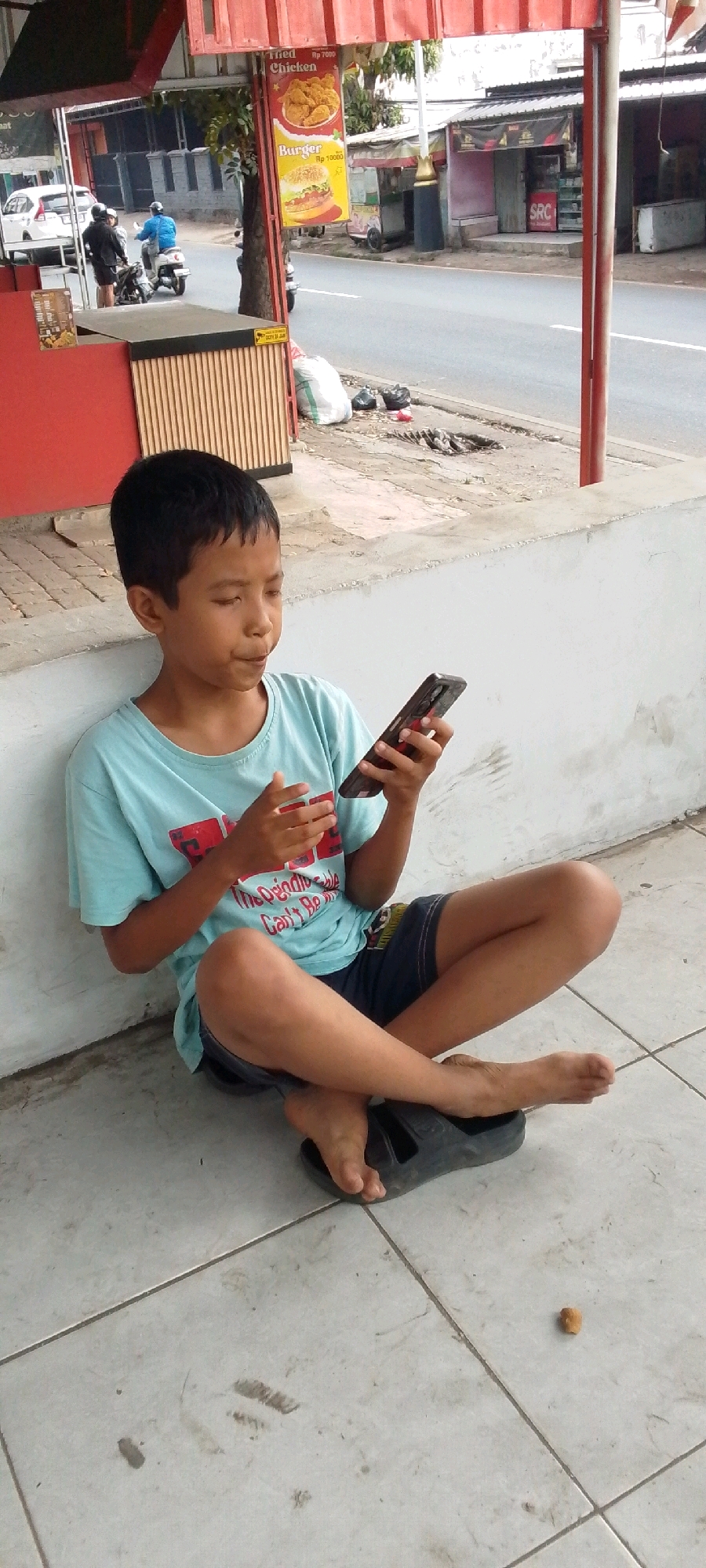 In Cilaku, Indonesia, a young boy with short, dark hair is sitting cross-legged on a tiled floor, leaning against a light-colored wall. He is wearing a light blue t-shirt with red text and dark shorts. The boy is holding a dark-colored smartphone in his hands, appearing to be actively engaged with it. His feet are bare, and one is resting on a black sandal. The environment suggests an outdoor setting, possibly near a street or a building. In the background, a street is visible with some businesses and a red and white awning overhead. A white car and two motorcycles are also present on the street. The ground is tiled, with some dirt and debris scattered around, including a small piece of brown material. A few trash bags are visible near the wall.