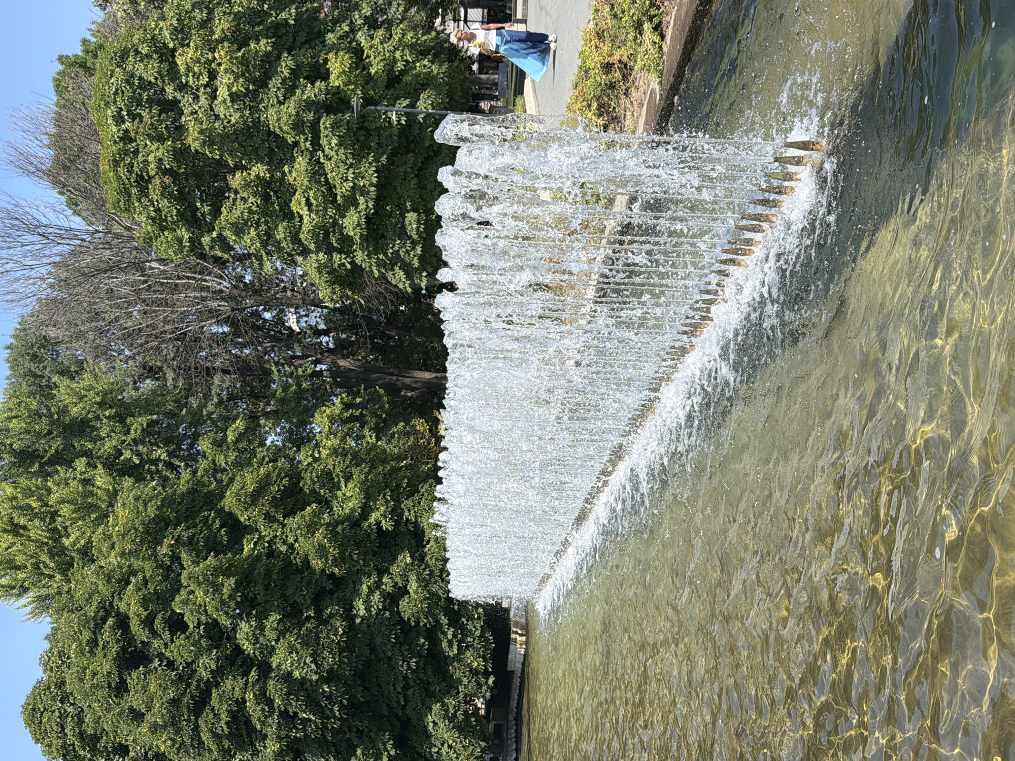 This image depicts a linear fountain with multiple water jets creating a curtain of water. The fountain is situated in a body of water with a textured surface, reflecting sunlight and creating dappled patterns. Behind the fountain, there is a paved walkway with a woman walking to the right. She is wearing a white top and a long, flowing blue skirt. Lush green trees form the background, with a clear blue sky above. The environment suggests a public park or outdoor recreational area, consistent with the Kaylaka Park in Pleven, Bulgaria, which features a large fountain and extensive green spaces. The scene is illuminated by natural daylight, indicating it was taken during the daytime.