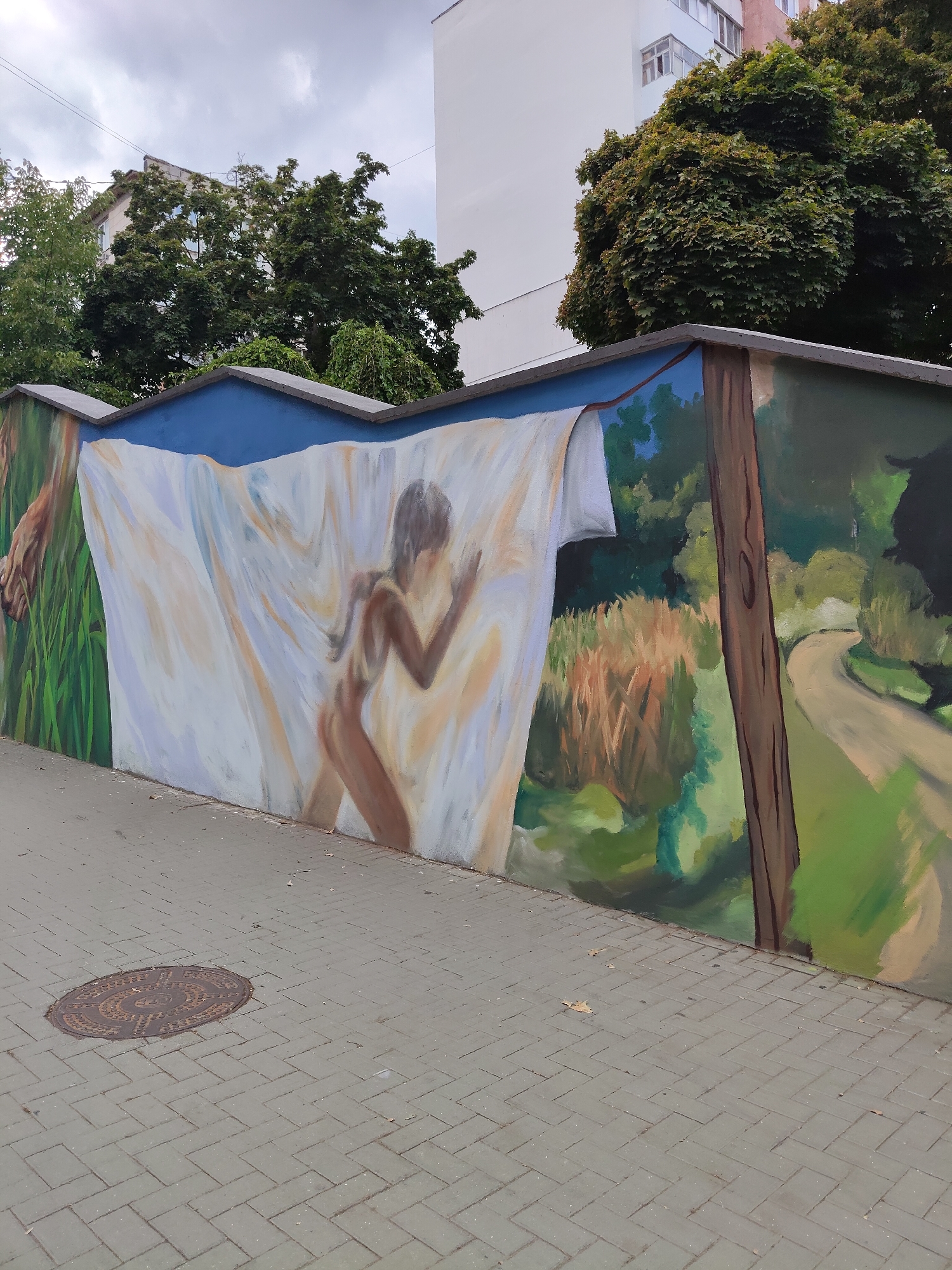 The image shows a mural painted on a concrete wall in Chișinău, Moldova. The mural depicts a scene with a figure partially obscured by a white sheet, appearing to be dancing or moving. To the left, a hand is visible amongst green grass. To the right of the figure, there is a landscape with trees, tall grass, and a winding path. A wooden pole is painted on the right side of the mural. The wall is located next to a paved walkway with a manhole cover. Buildings and trees are visible in the background. The sky is overcast.