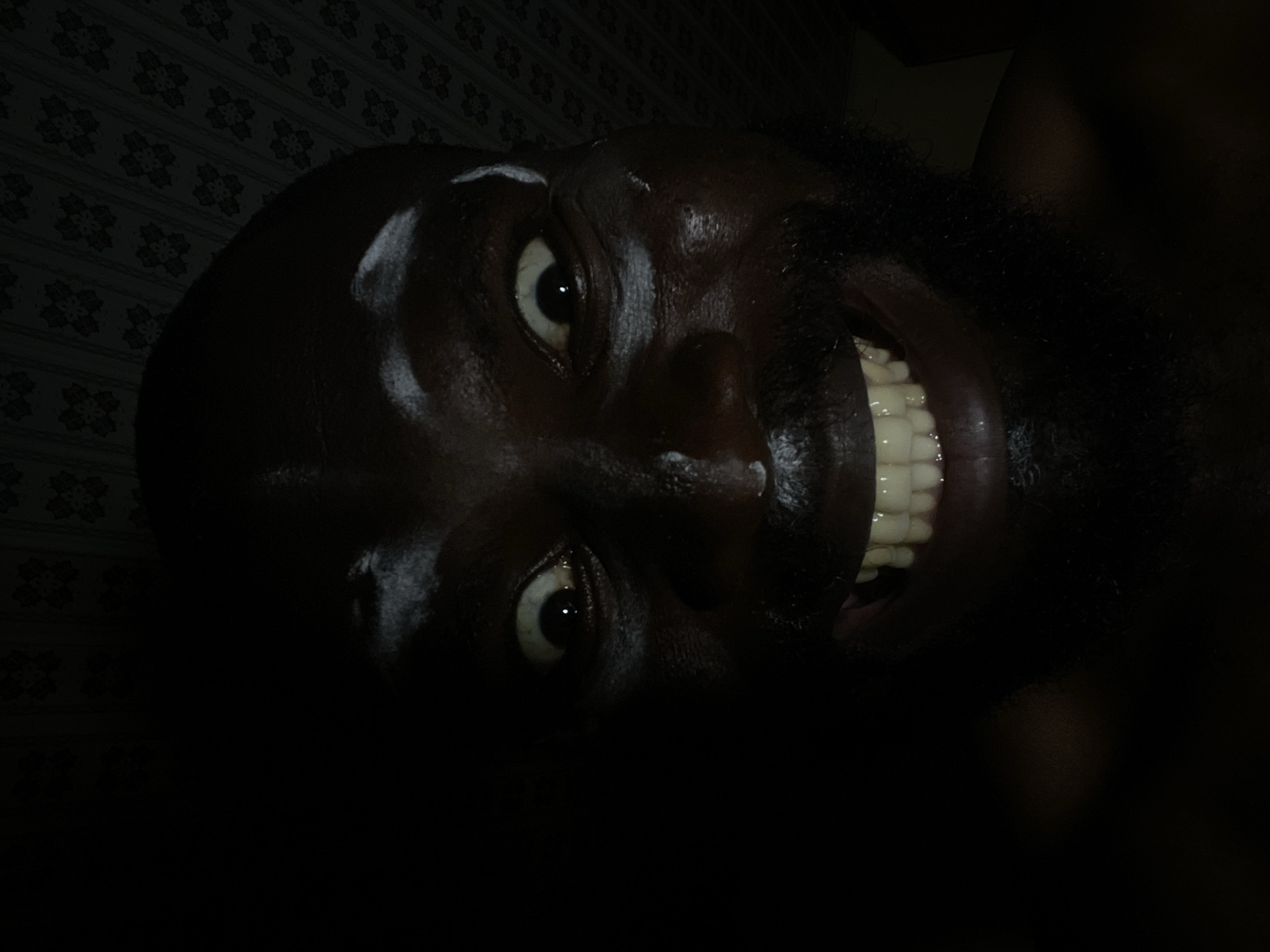 A dark, close-up image of a Black man's face is presented. His head is tilted to the side, and he is smiling widely, exposing a set of white teeth. The top of his head appears bald, with some reflected light. The background is dark and appears to be a wall with a faint pattern. The location, Gura Top, Nigeria, is not discernible within the image.