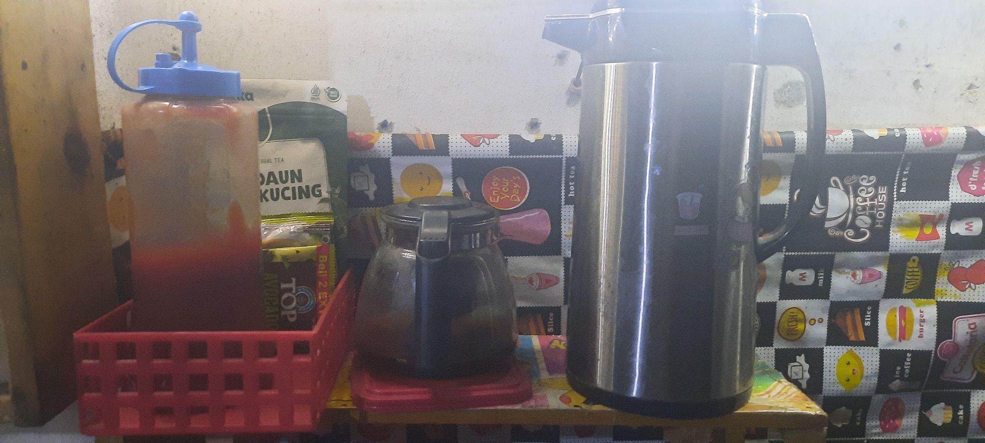 The image depicts a close-up, eye-level view of a shelf in what appears to be a kitchen or food stall. On the left, a clear plastic squeeze bottle with a blue cap is filled with a red liquid, likely a sauce or condiment, and sits in a red plastic basket. To the right of the bottle are several packets of tea, including one labeled "DAUN KUCING" (Cat Leaf) and another with "TOP AVOCADO". In the center, a dark glass teapot with a silver lid is positioned on a red coaster. Further to the right is a large, tall, silver thermal carafe. The shelf is covered with a black and white checkered fabric featuring various food-related illustrations and text, including "Coffee House", "hot tea", "burger", "cake", and "lemon". The background wall is a textured, off-white color. The lighting suggests an indoor setting with a bright light source reflecting off the silver carafe. The scene conveys a sense of preparation or serving of beverages and condiments. The location is specified as Jatitengah, Indonesia.