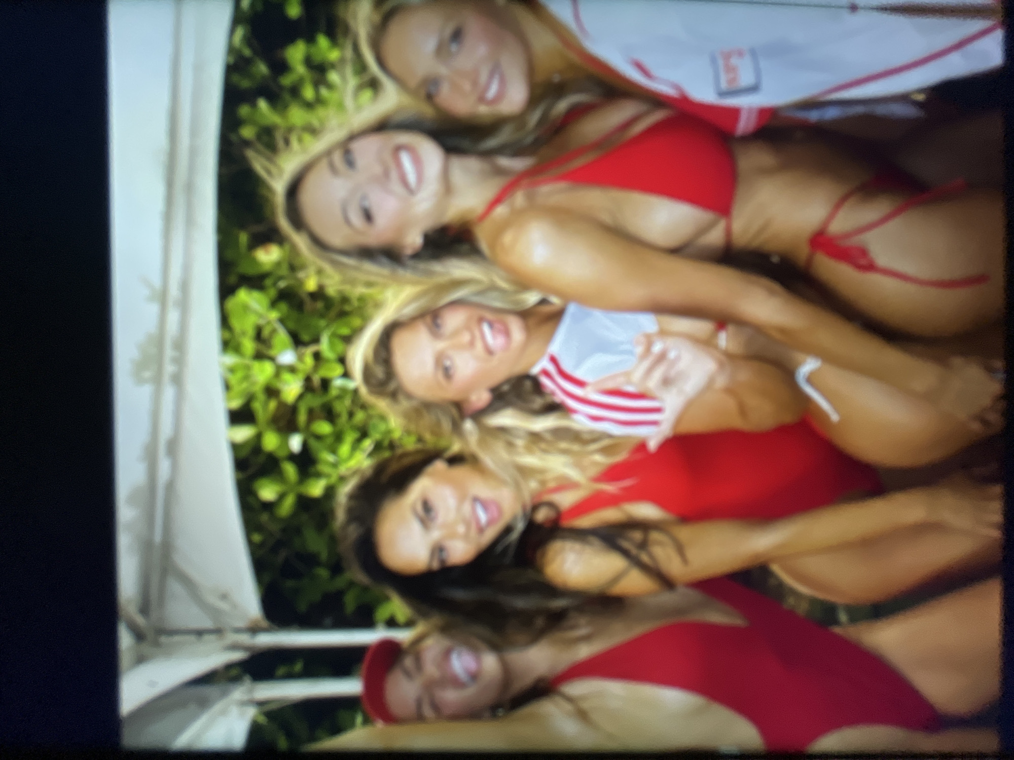 A group of five young women, all wearing red swimwear, pose for a photo in an outdoor setting. The women are smiling and appear to be in good spirits. One woman on the far left wears a red cap. The background consists of lush green foliage, suggesting a tropical or garden environment. A white tent-like structure is visible overhead, possibly indicating a party or event. There is no indication of the city of Sofia, Bulgaria, in the image.