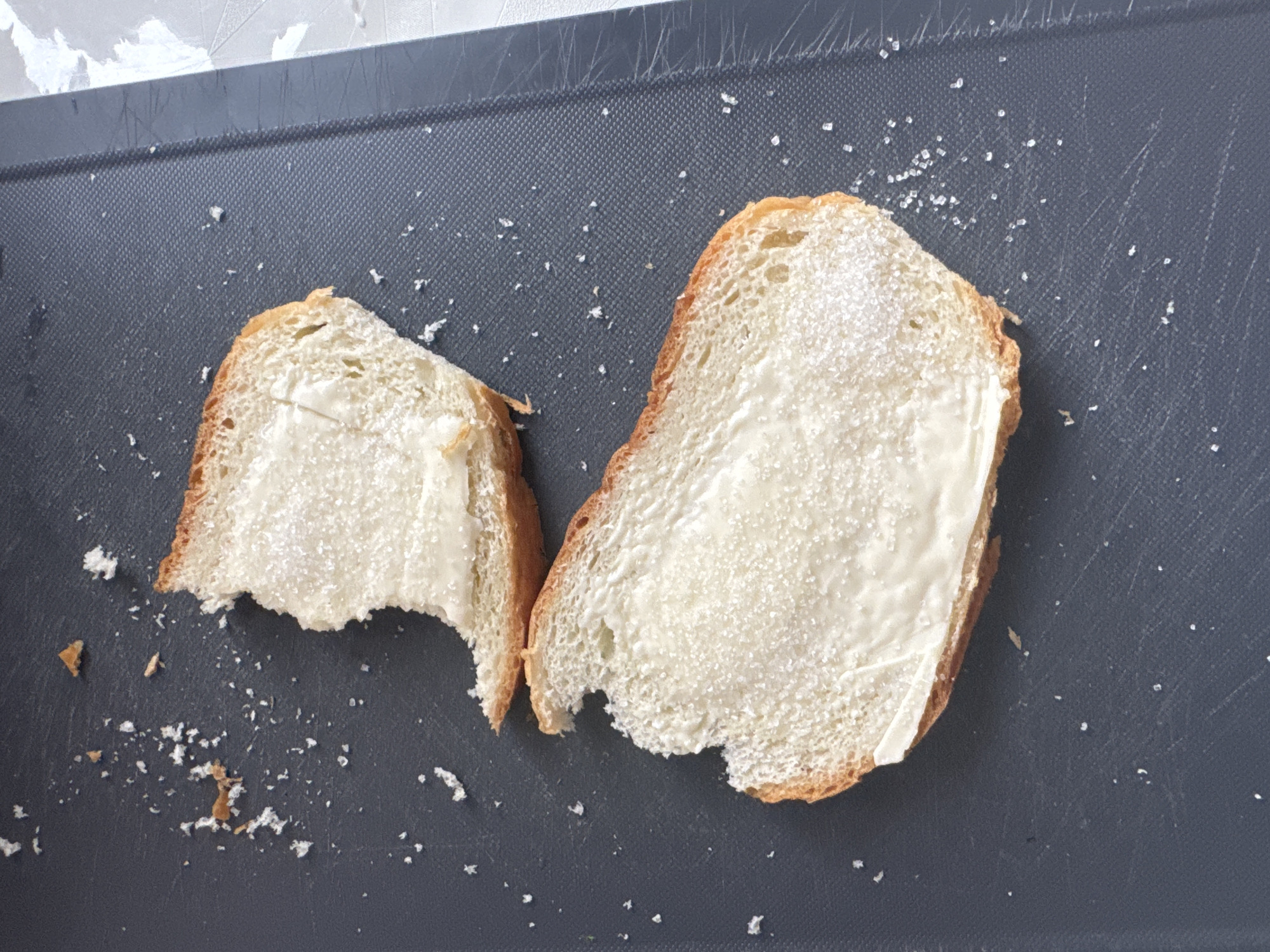 On a gray cutting board are two slices of white bread. Each slice is spread with a white substance, possibly butter or margarine, and sprinkled with a granular substance, likely sugar. One slice is partially eaten, with a bite missing. Breadcrumbs and sugar granules are scattered around the bread on the cutting board. The background includes a glimpse of a gray and white surface, potentially a countertop or wall. The location of Vesyoloye, Russia is not discernible from the image.