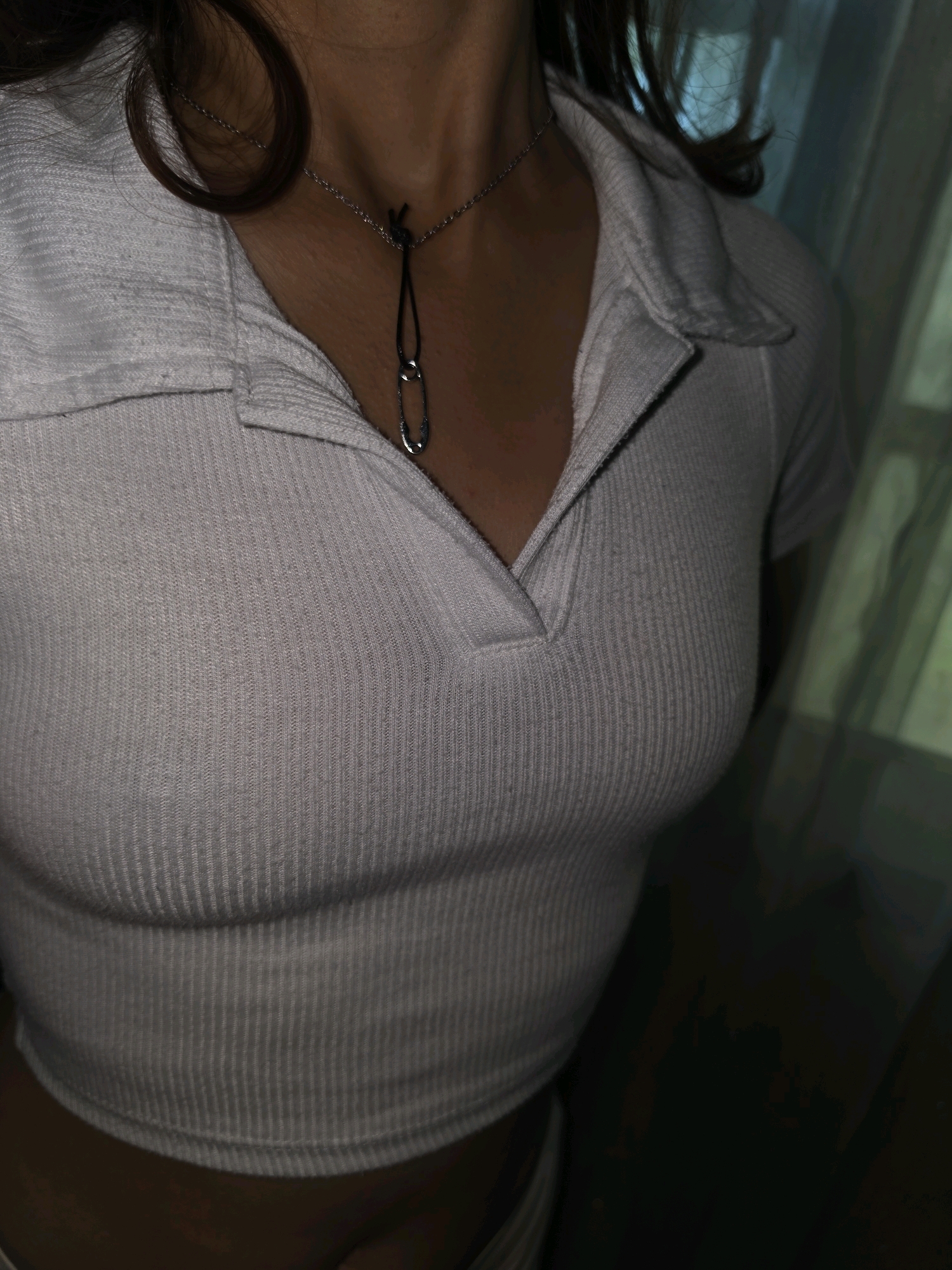 The image is a close-up, waist-up shot of a person wearing a white, ribbed, short-sleeved polo shirt. A silver chain necklace with a black cord and a silver safety pin pendant is visible around the person's neck. The background is dark and out of focus, with some hints of green in what appears to be a curtain or blinds. There is no indication of Pintomakmur, Indonesia, or any urban environment. The image appears to be taken indoors, with the subject in focus and the background blurred.