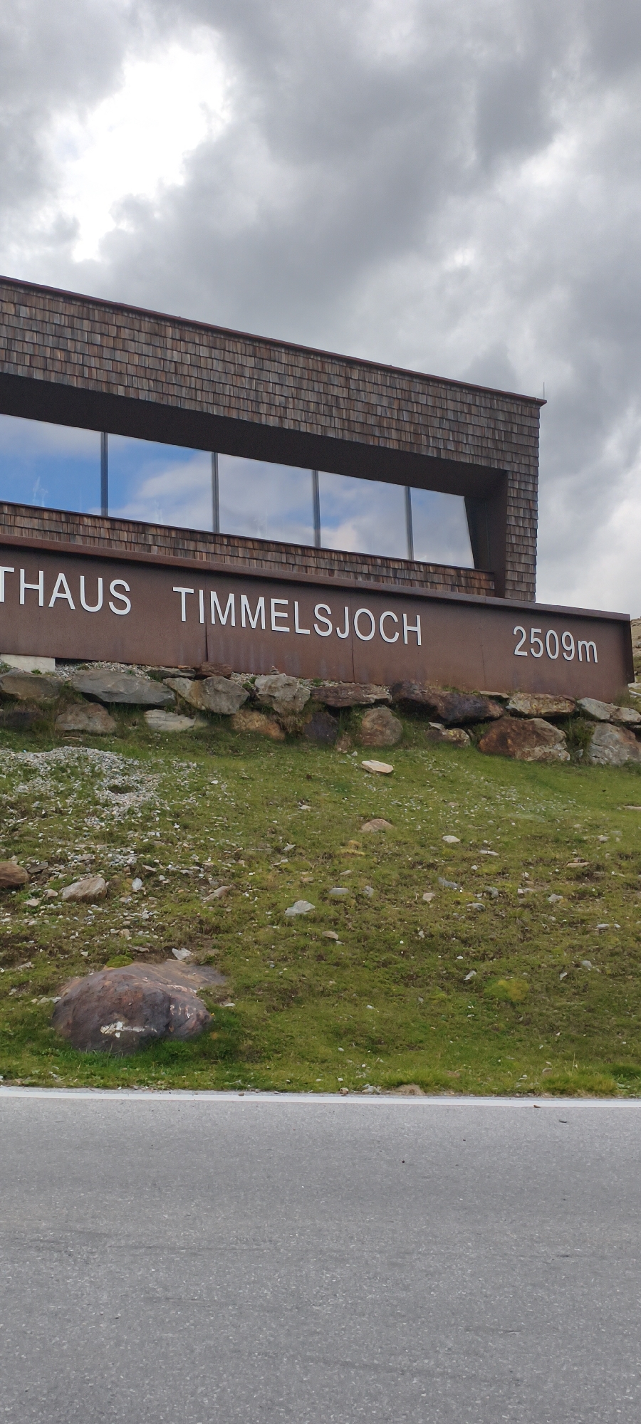 The image depicts the "Timmelsjoch" building, a modern structure with a wood-shingled facade and large windows reflecting a cloudy sky. Below the windows, a rusted metal sign displays the text "THAUS TIMMELSJOCH" in white lettering on the left, and "2509m" on the right. The building is situated on a grassy slope with scattered rocks. The foreground shows an asphalt road with a white line marking its edge. The image is taken from a low angle, looking up at the building. There are no people visible in the scene. The Timmelsjoch is a mountain pass in the Ötztal Alps, connecting the Austrian state of Tyrol with the Italian province of South Tyrol. Sölden is a municipality in the Ötztal valley.