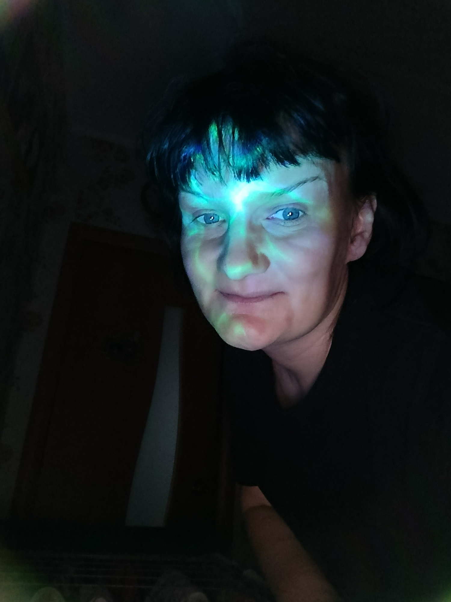 A woman with short, dark hair and blue eyes is positioned in a dark room, illuminated by a projected green and blue light that covers her face. She is wearing a black shirt. In the background, a dark wooden door is visible on the left side of the frame. The scene appears to be indoors, with a patterned wall faintly visible behind the door.
