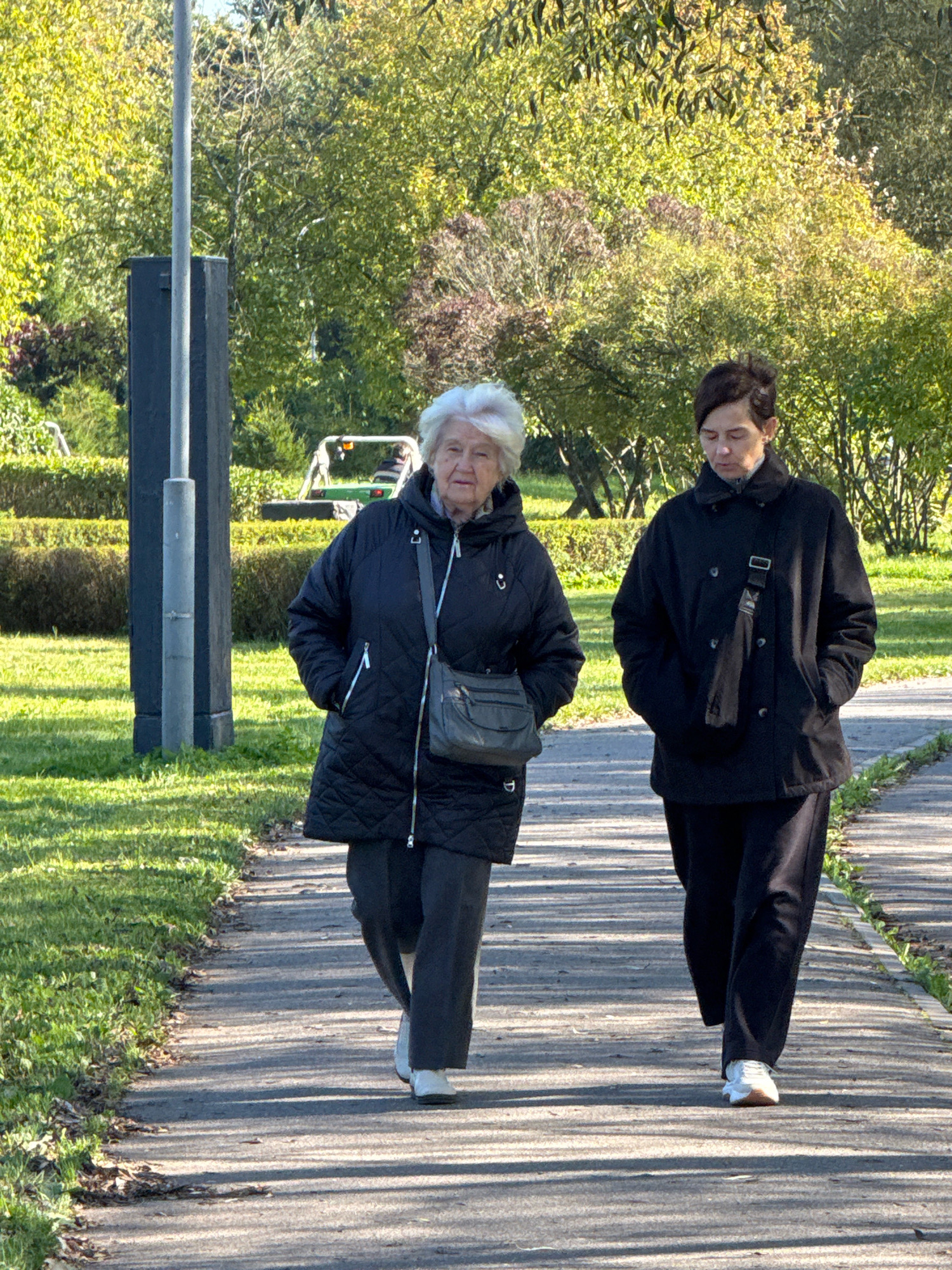 Two women are walking on a paved path in a park. The woman on the left has white hair and is wearing a black quilted jacket and dark pants. She is carrying a dark grey crossbody bag. The woman on the right has dark hair and is wearing a black coat and dark pants. She has a black strap across her chest, with a dark bag hanging on her hip. The path is lined with green grass and trees in the background. A lamppost is visible on the left side of the image, near a dark, rectangular structure. In the distance, a green tractor is operating. The scene suggests a sunny autumn day. This scene could be located in any park in Saint Petersburg, Russia, as parks with paved paths, grass, trees, and public amenities like lampposts are common.