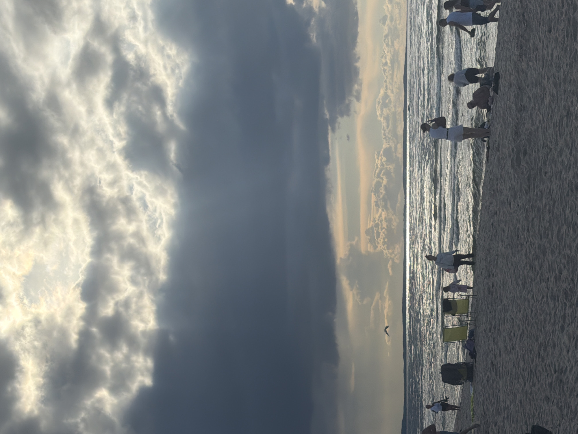 The image captures a beach scene in Międzyzdroje, Poland, under a dramatic sky filled with dark, textured clouds interspersed with bright, sunlit areas. The sun's rays break through the clouds, creating a luminous effect over the water. The sea is moderately choppy, reflecting the light and creating shimmering patterns on its surface.
On the sandy beach in the foreground, several people are visible. On the left, a person is partially visible, appearing to be walking. Further to the right, a woman stands near the water's edge, holding what appears to be a phone to her ear. Next to her, a person is sitting on the sand. A group of people are gathered closer to the water, including a man and two women. One of the women is carrying a bag over her shoulder. Further to the right, a couple walks along the shoreline. In the middle ground, two empty yellow beach chairs are set up, with a bag and some items lying on the sand near them. A child in a light-colored garment stands between the chairs and the water. Another woman stands to the right of the chairs, holding a bag.
A bird is captured in flight over the water, silhouetted against the bright sky. The overall atmosphere suggests a late afternoon or early evening at the beach, with the weather adding a sense of drama to the scene. The location is identifiable as Międzyzdroje due to the characteristic coastal landscape and the prevalent cloudy, yet bright, sky often seen in this Baltic Sea resort town during certain times of the year.
