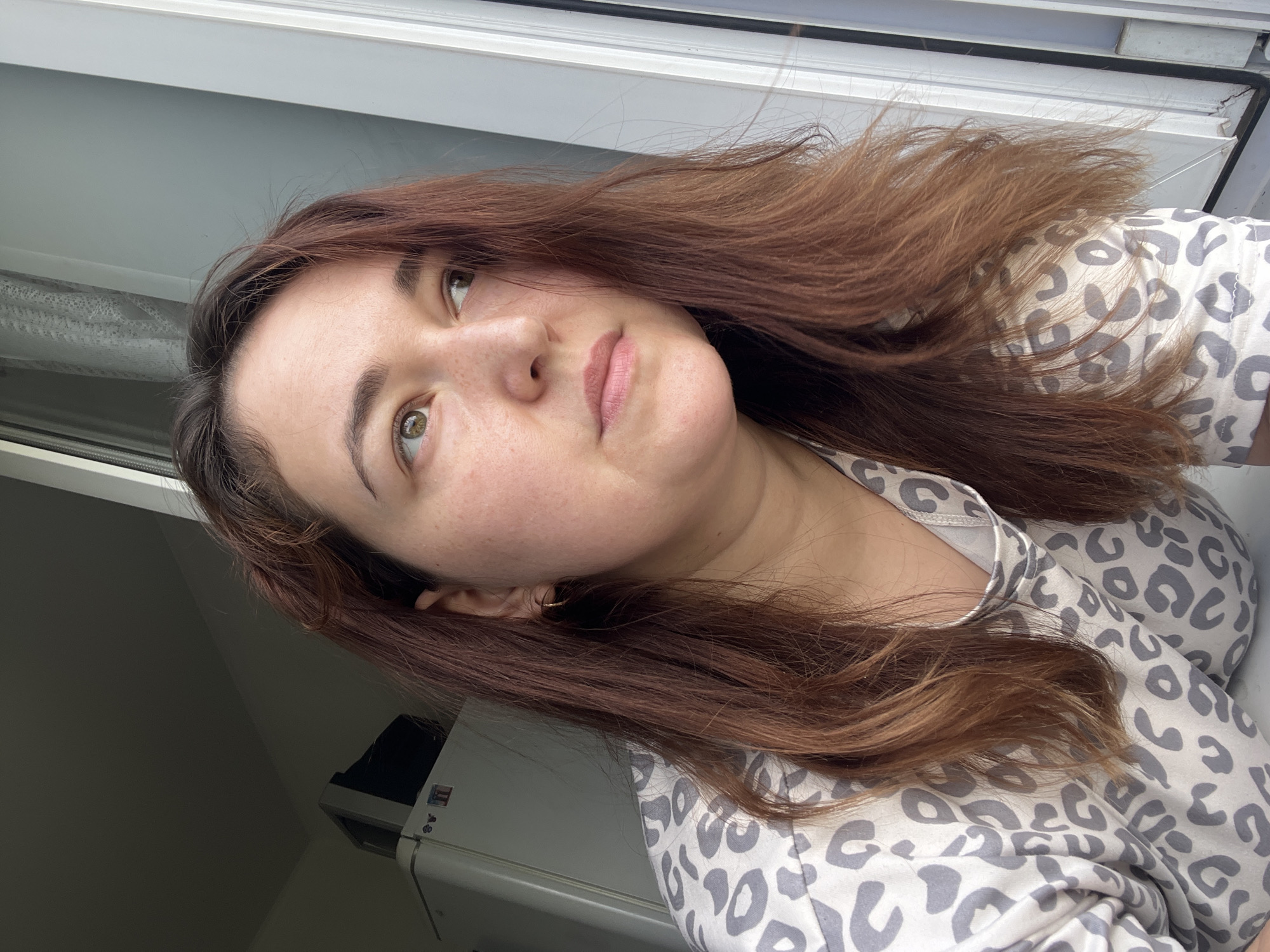 A woman with long, auburn hair is shown in a close-up, profile view, looking out a window. She is wearing a light-colored pajama top with a grey leopard print. In the background, a white refrigerator is visible. The scene appears to be indoors, with natural light coming from the window. The location is not specified in the image.