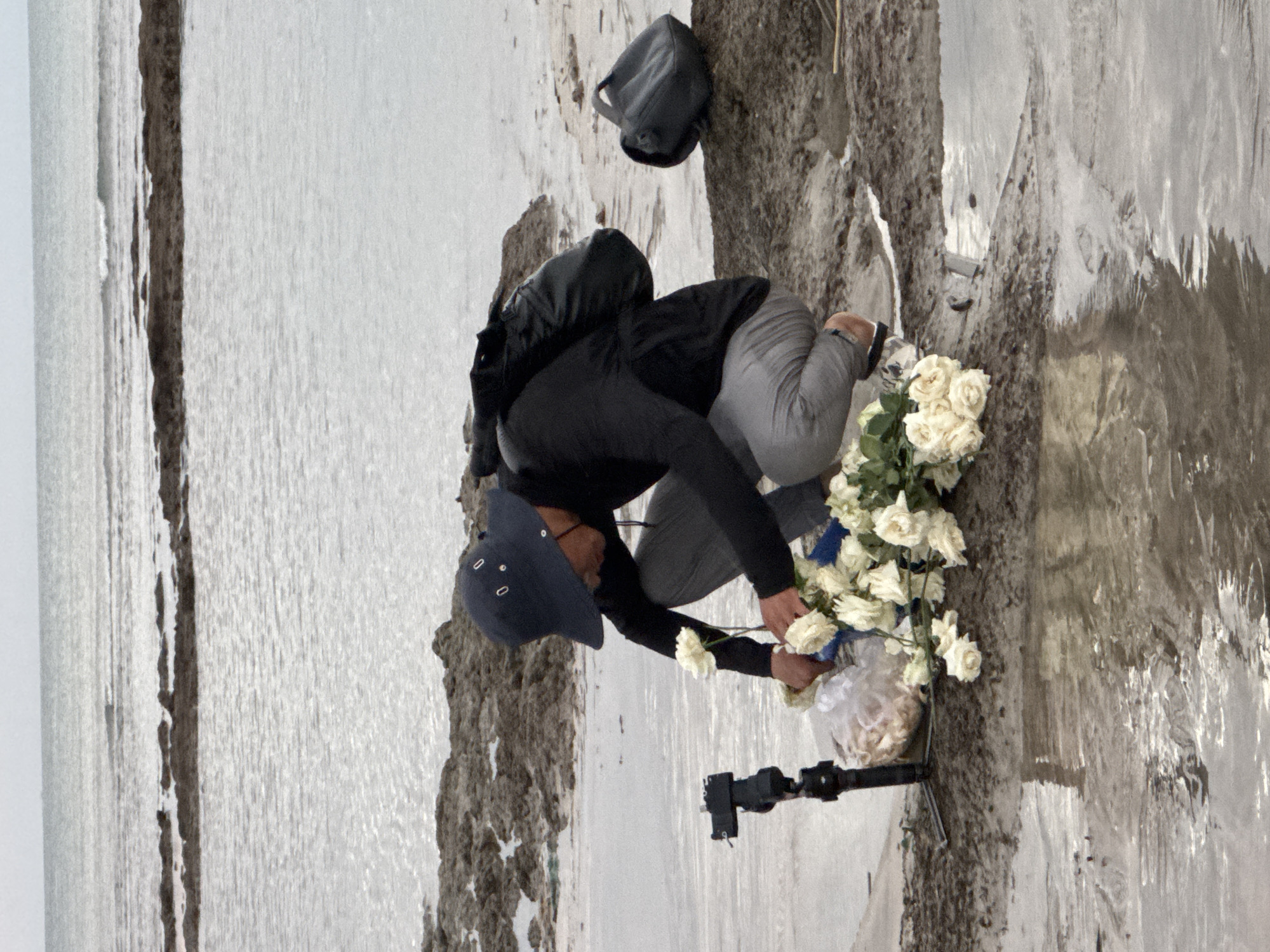 A person wearing a dark blue bucket hat and a dark long-sleeved shirt is crouching on a muddy shore near the ocean. They are placing a bouquet of white roses on the ground. In front of them, to the left, is a camera on a tripod. To the right, further back, is a dark bag. The ocean is visible in the background with rocky formations extending into the water. The sky is overcast. The scene suggests a memorial or tribute. The location is Kuta Selatan, Indonesia.
