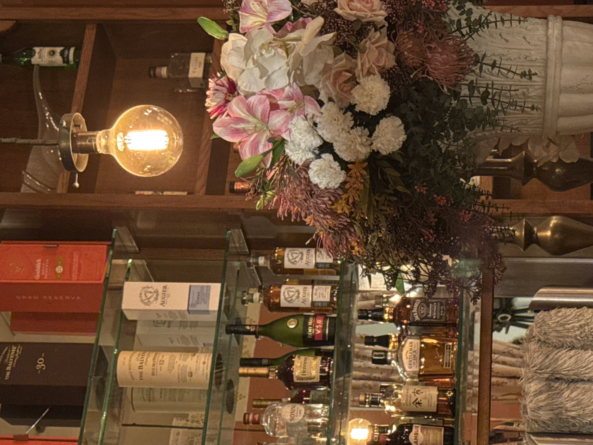 The image shows a bar shelf in what appears to be a restaurant or lounge, possibly located in Tel-Aviv, Israel (due to the Hebrew writing on one of the bottles, “余市”). The shelves are made of glass, displaying a variety of alcoholic beverages including whisky (e.g., The Balvenie, Jack Daniel's), cognac (e.g., Hennessy, Remy Martin), and liquor (e.g., Nonino Ambur). Brands like "Augier" are also visible. The bottles are arranged in neat rows. The shelves are wooden, and the wall behind them is also wood.
A floral arrangement with pink, white, and green flowers and greenery is centered above the shelves, partially obscuring some of the bottles. An antique-style lightbulb is located above and to the left of the floral arrangement. The bulb is exposed with a visible filament.
A piece of furniture, possibly a sofa or chair with a beige, textured surface, is visible on the right side of the image.