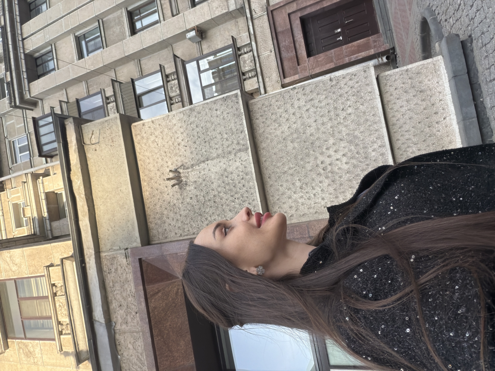 A woman is looking up while standing next to a building in Saint Petersburg, Russia. The building has a façade made of concrete and stone, and several windows are visible. The woman has long brown hair and is wearing a black sequined jacket. The sky is cloudy and the lighting is soft.