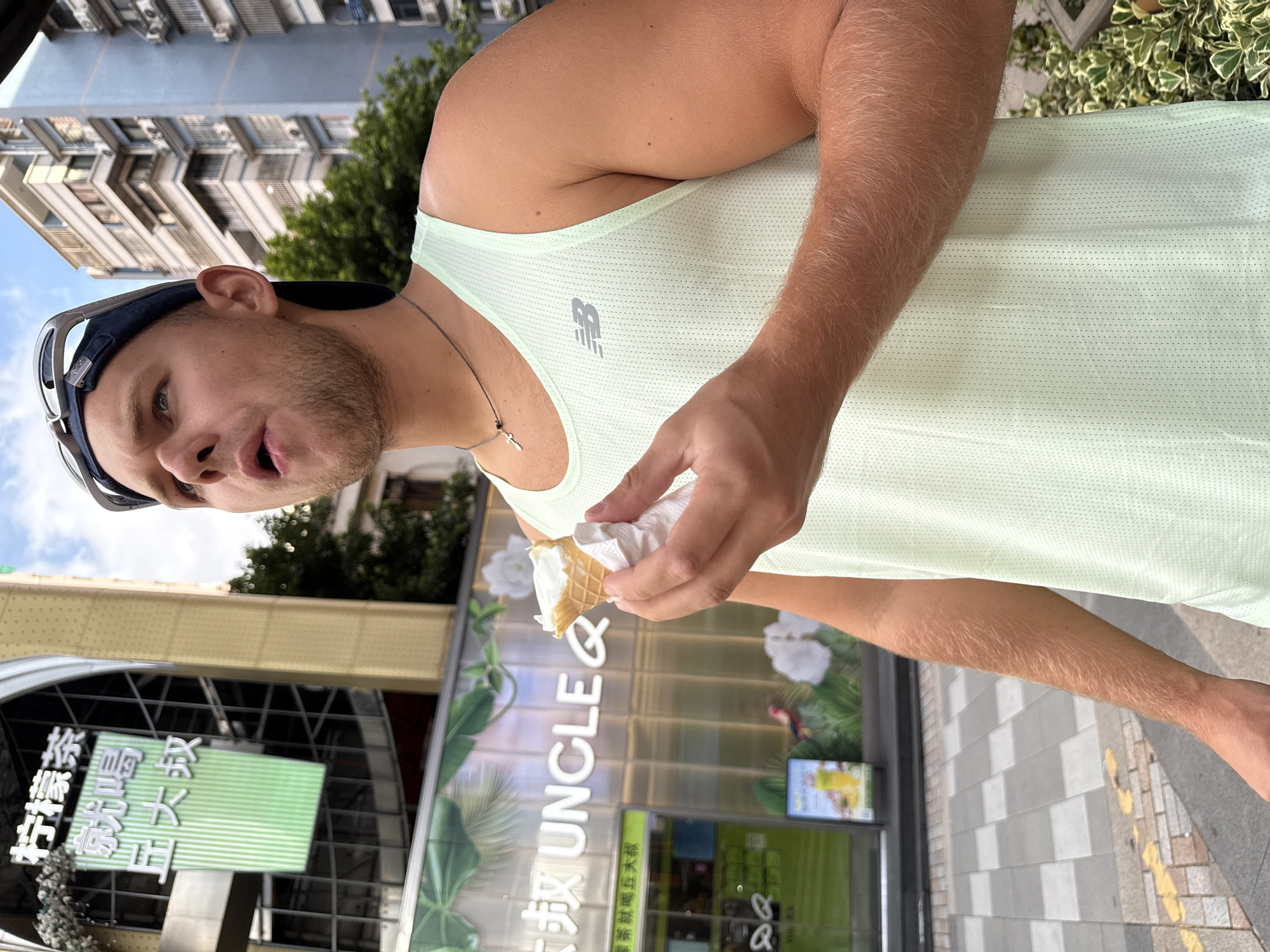In the Haizhu District of China, a young Caucasian male is captured mid-action, holding a partially eaten ice cream cone wrapped in a white napkin in his right hand. He wears a light mint-green tank top with a New Balance logo, a dark baseball cap with sunglasses perched on top, and a thin chain necklace with a cross pendant. The man appears to be looking up and to his left, his mouth slightly open as if about to take another bite of the ice cream.
The background reveals a bustling urban environment. To the left, a building with prominent Chinese signage, "柠檬茶" (Ningmeng Cha) and "京喝 丘大叔" (Jing He Qiu Da Shu), stands tall, adorned with decorative white elements. Below this, a glass-fronted establishment with the name "UNCLE Q" in large white letters is visible, featuring lush green foliage and floral graphics on its facade. A menu board displaying a yellow drink is positioned near the entrance. The ground is paved with a checkered pattern of gray and white tiles, with yellow painted lines indicating a pedestrian crossing. To the right, a tall, light blue apartment building with numerous windows and air conditioning units dominates the skyline, surrounded by verdant trees and shrubbery. The sky above is a bright blue with scattered clouds.