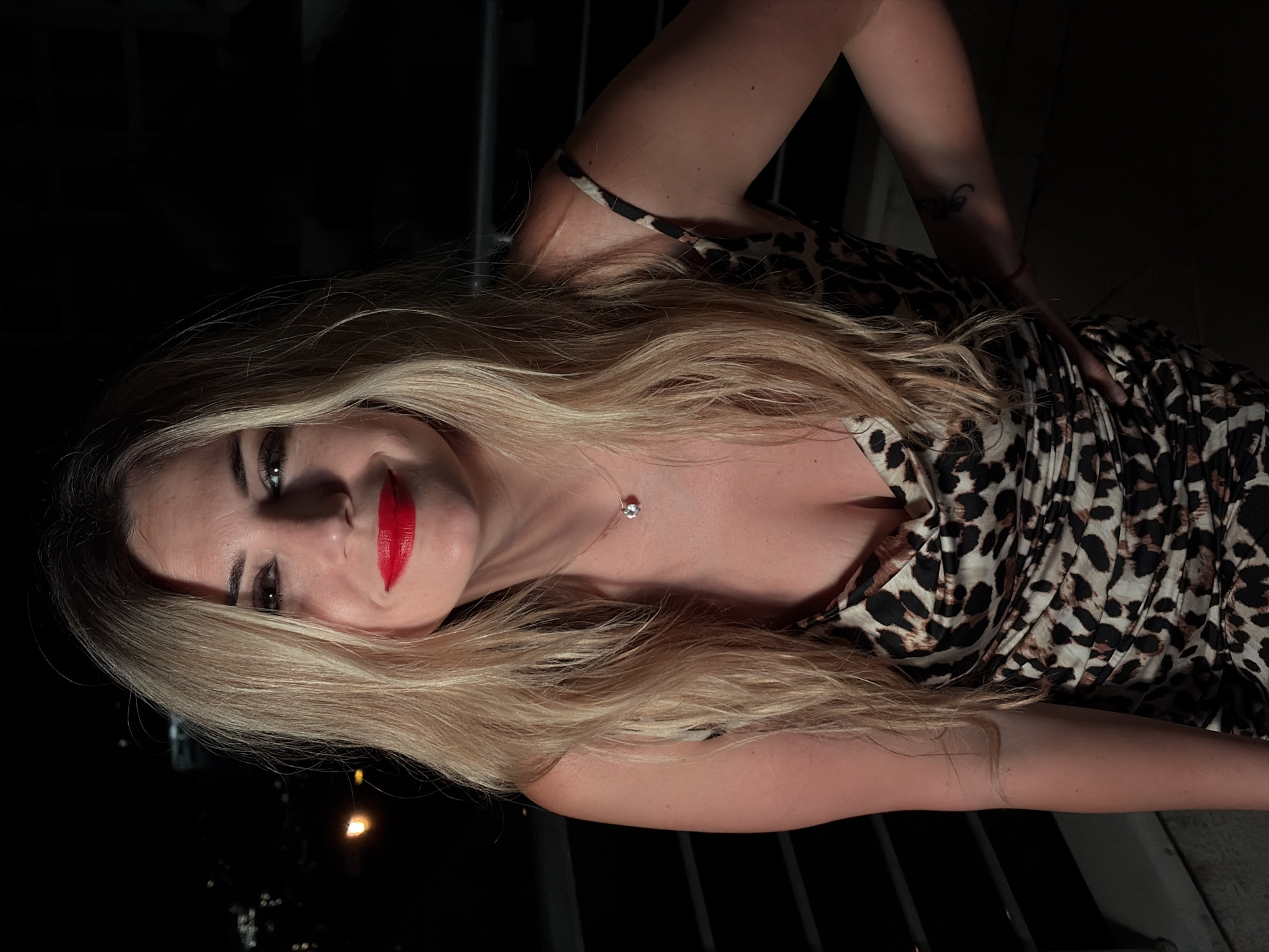 A woman with long blonde hair and bright red lipstick is pictured in a close-up shot. She is wearing a leopard-print dress with a cowl neck and a delicate necklace with a small pendant. Her left arm is bent, with her hand resting on her hip. A faint tattoo is visible on her right forearm. The background is dark, with blurred lights suggesting an urban nighttime setting, possibly on a balcony in Budva, Montenegro.