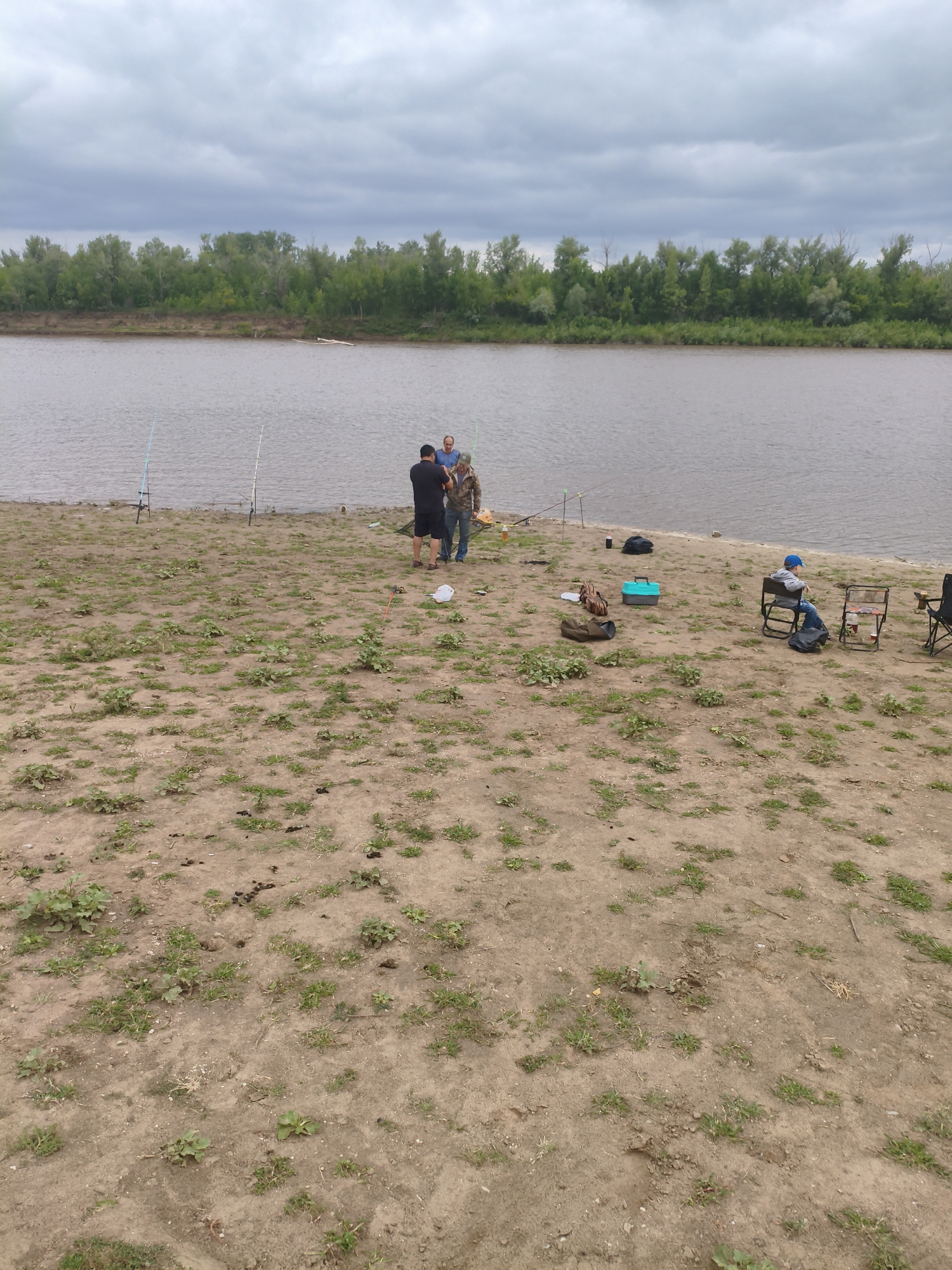 The image depicts a natural landscape with a river and a tree line under a cloudy sky. Several individuals are present on the sandy bank of the river, engaged in fishing activities. Three fishing rods are visible, planted in the ground near the water's edge. Three men are standing near the river, one in a black t-shirt and shorts, another in a blue shirt, and a third in a camouflage jacket. A young person wearing a blue cap and jeans is seated in a folding chair further down the bank. Various fishing gear and bags are scattered on the ground, including a blue tackle box. The environment is characterized by dry, sandy soil with patches of sparse vegetation. The location is identified as the Ақсуат ауылдық округі in Kazakhstan.