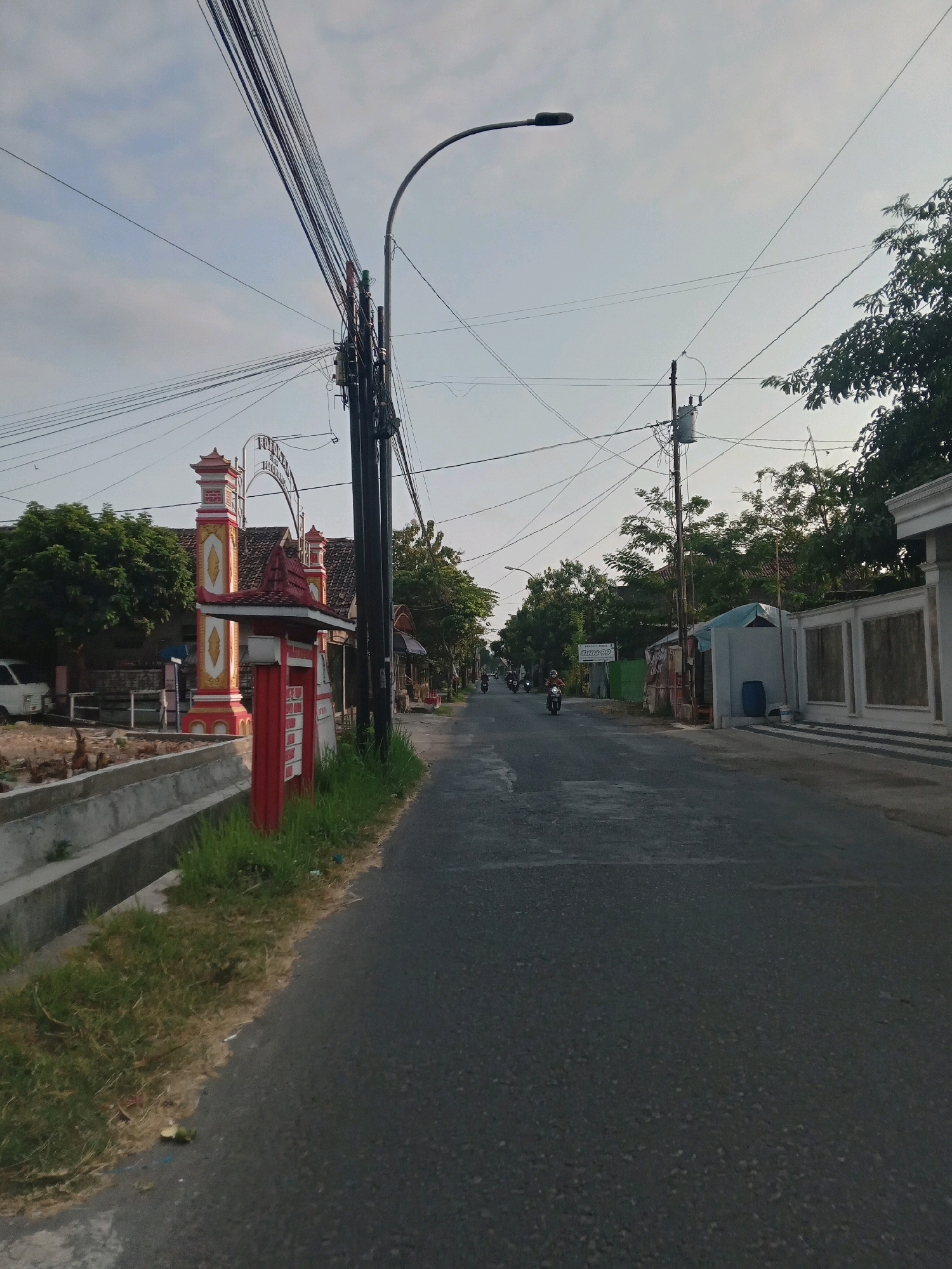 The image depicts a street scene in Banguntapan, Indonesia, under a bright sky with scattered clouds. The perspective is from the middle of an asphalt road looking forward. On the left side of the road, there is a red and yellow decorative gateway structure with a tiled roof, possibly marking an entrance to a residential area or a specific building. Adjacent to this structure is a concrete drainage ditch with some grass growing along its edges. A white van is parked to the left, behind the gateway. Further down the road, on the left, are trees and some buildings.
On the right side of the road, there are more buildings, including one with a white wall and decorative panels. A blue plastic barrel is visible next to a small structure with a tarp roof. Overhead, a network of black electrical wires is prominent, supported by utility poles, one of which has a transformer. A street lamp is visible on the left side of the road, near the gateway.
Several motorcycles are traveling on the road, with riders visible on some. The overall environment appears to be a suburban or semi-urban setting with signs of daily life, including infrastructure for utilities and transportation. The lighting suggests it is either early morning or late afternoon.