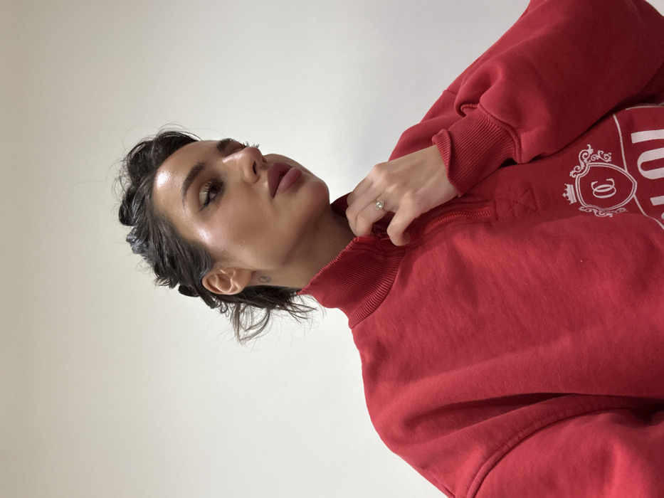 A woman with slicked-back dark hair is seen from a low angle, glancing upward with a serious expression. She wears a bright red, collared sweatshirt with a white crest design. Her hand is near the collar, and a ring is visible on her finger. Her skin is luminous, and the background is a plain white wall. The photo has been rotated slightly clockwise.