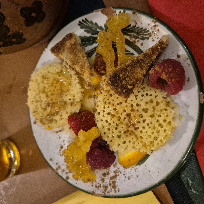 A decorative plate displays a dessert. There are two types of spongy cakes, one with a porous texture. Toasted triangular bread slices are placed between the cakes. Red raspberries and translucent yellow candied pieces adorn the plate. A sprinkle of brown powder, possibly cinnamon, is dusted across the dessert and the plate's surface. The plate has green trim and painted foliage.