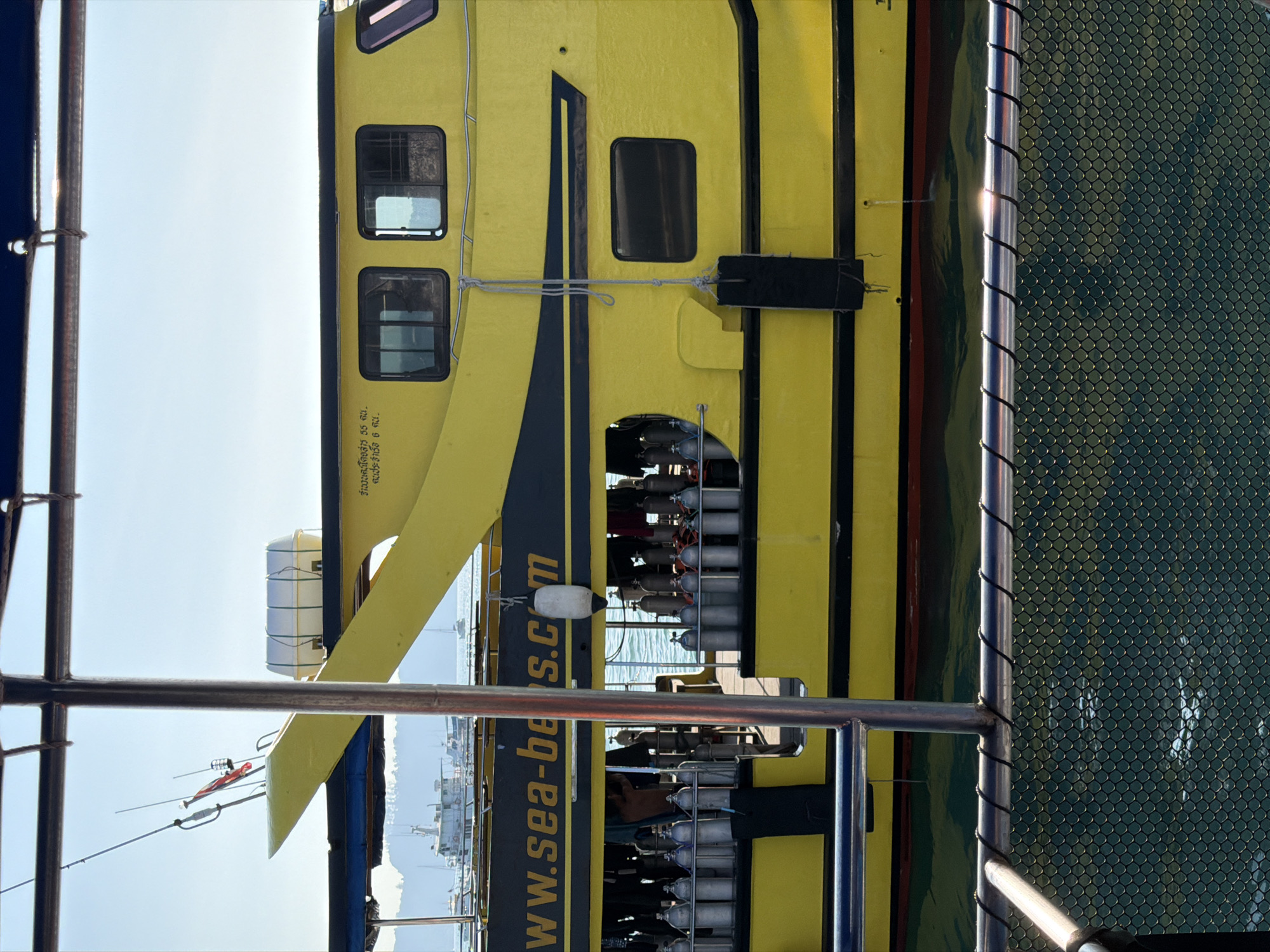 The image displays the port side of a large dive boat, predominantly yellow with black accent stripes, moored in the waters of Rawai, Thailand. The partially visible website address "www.sea-be s.c m" is stenciled in yellow on a black stripe along the hull. Thai script is visible on the bow structure.
**Subjects & Objects:**
* **Boat:** The central vessel features a forward cabin with multiple windows. A white oval life raft container is mounted on the upper forward deck. Antennas and a flag are affixed to a mast-like structure at the bow.
* **Diving Equipment:** An open deck area midship contains numerous silver compressed air diving tanks, arranged in multiple rows.
* **Fenders:** A white cylindrical fender and a black rectangular fender hang from the boat's side, secured by ropes.
* **Waterline:** A red antifouling paint line is visible along the hull's submerged portion.
* **Foreground Elements:** The image is framed by dark metallic railings and a diamond-pattern mesh fence, indicating the perspective from another vessel or a dock. A prominent vertical metal support pole is centered in the foreground.
**People:**
* At least two human figures are partially visible within the open deck area among the diving tanks, suggesting crew or passengers. One figure's upper torso and head are discernible on the port side, while another is further inside the vessel.
**Environment & Background:**
* The water is a calm, deep teal or green color, with slight surface ripples.
* The background features a clear, bright blue sky above distant, lush green, tree-covered land or mountains.
* Several other boats, including a larger white vessel and smaller craft, are visible in the mid-distance on the water.