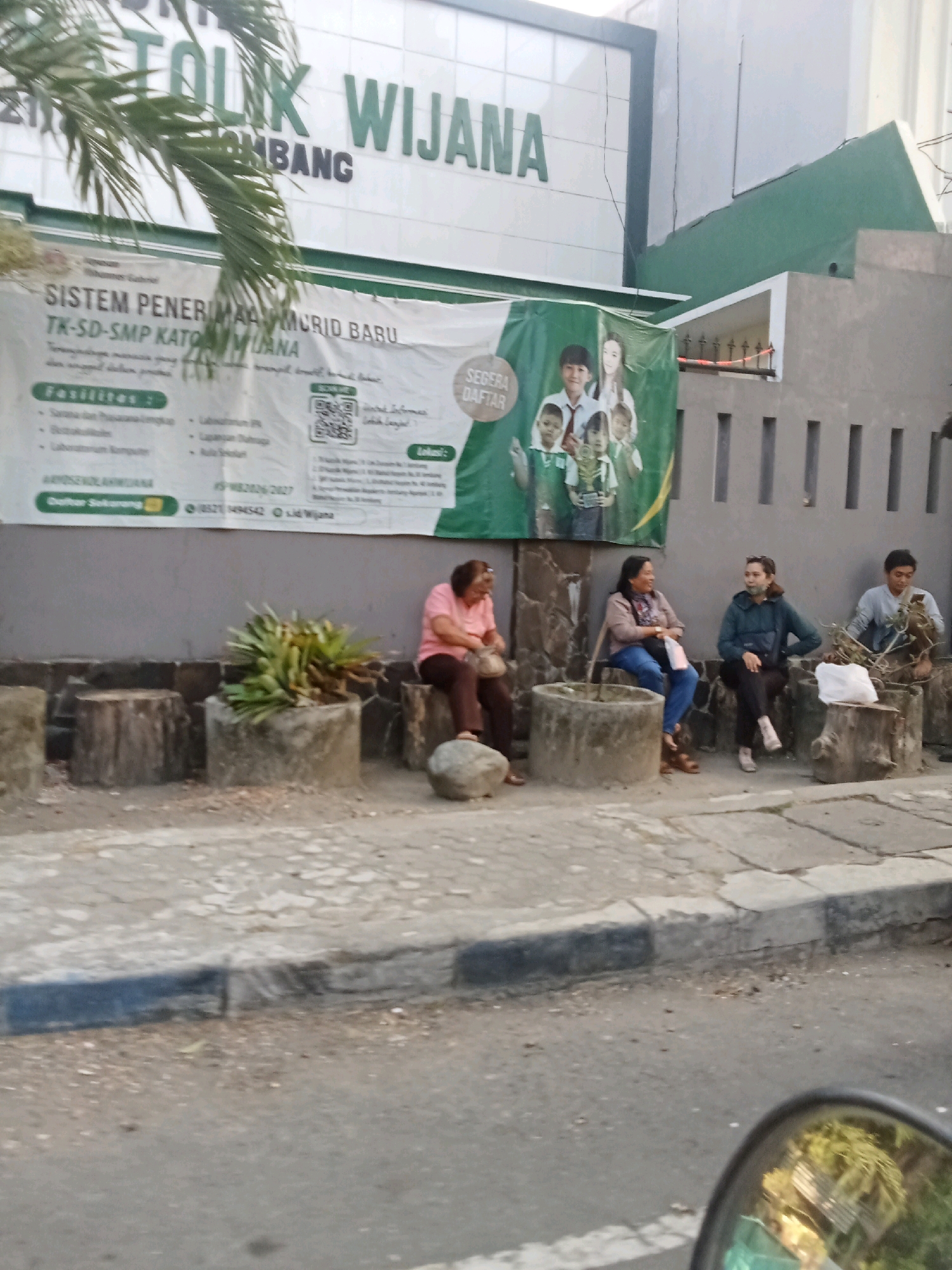 A group of people are sitting on concrete stumps and cylindrical planters in front of a building. The building has a sign that reads "TOLIK WIJANA" and also a banner advertising "SISTEM PENERIMAAN MURID BARU TK-SD-SMP KATOLIK WIJANA". The banner also lists facilities such as "Sarana dan Prasarana Lengkap", "Perpustakaan", "Laboratorium Komputer", "Laboratorium IPA", and "Lapangan Olahraga". On the right side of the banner, there is a QR code and contact information, including a phone number and an email address. The people are casually dressed and appear to be waiting or resting. The environment suggests an urban setting, likely in Jombang, Indonesia, given the context of the building's sign. The people are not interacting with each other in any significant way, and the scene is calm and uneventful. The time of day appears to be late afternoon or early evening, judging by the lighting conditions.