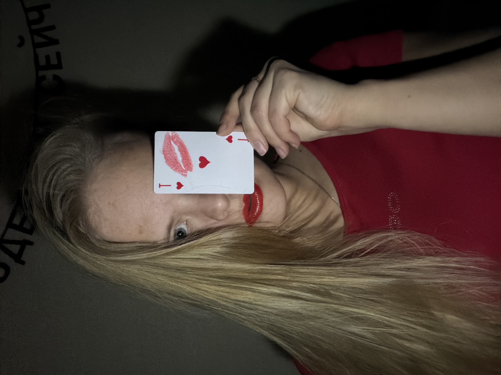 A woman with long, blonde hair is lying down, holding a playing card in front of her face. The card is the Ace of Hearts, with a lipstick mark on it. She is wearing a red shirt and has red lipstick on. The background is dark and out of focus, with some indistinct Russian text. The location is possibly Izhevsk, Russia, based on the text.