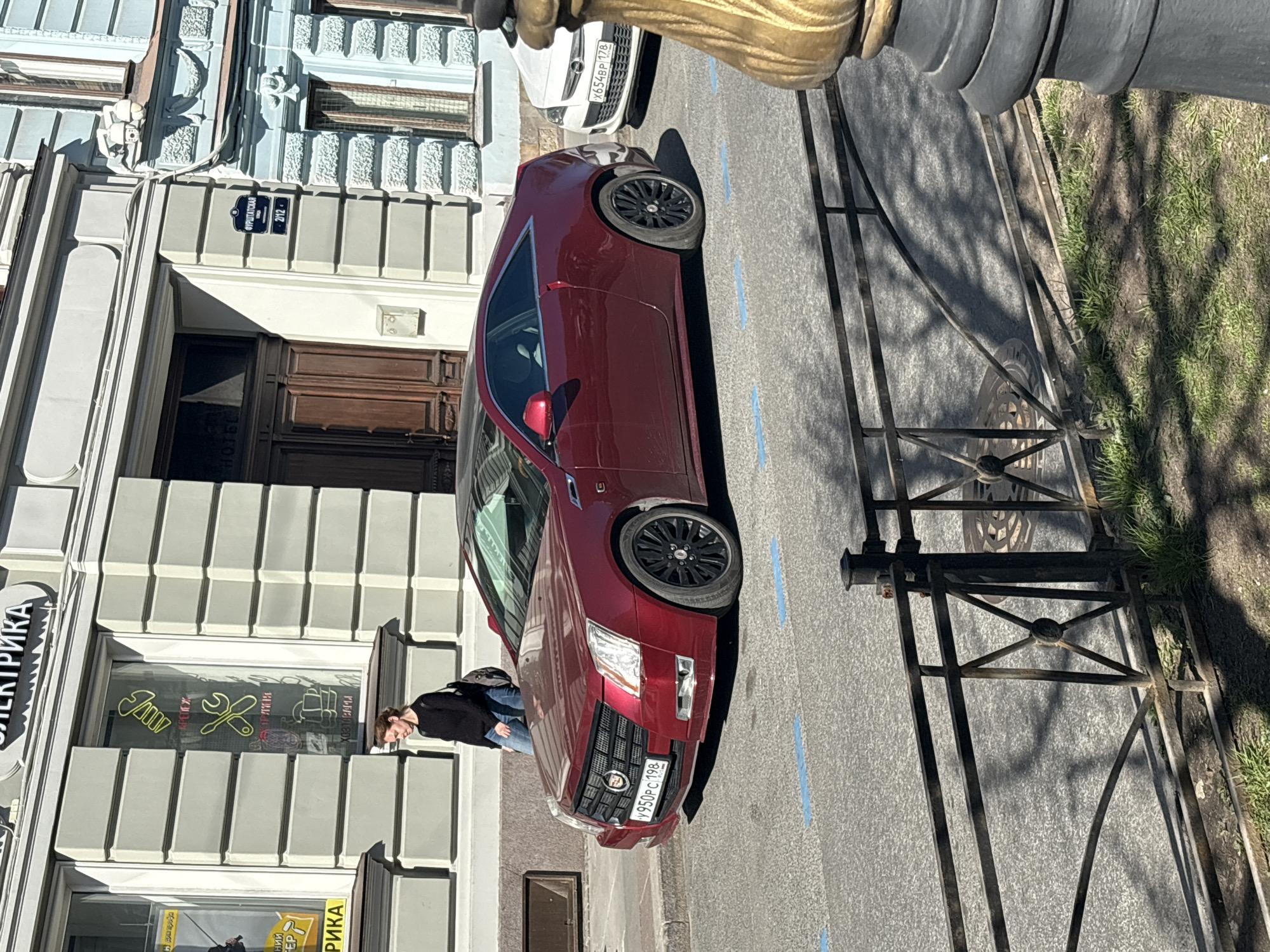 A daytime street scene in Saint Petersburg, Russia, shows a maroon Cadillac parked along the curb next to a sidewalk. A sign on the building reads "Фурштатская 2/12". A person is standing next to the front of the vehicle and appears to be looking at something in a window of a business in the building. The business window has "ЭЛЕКТРИКА" written vertically on the left. A white car is partially visible in the background. The street is marked with blue parking lines. A section of iron fence is visible along the sidewalk, and grass is visible behind the fence.
