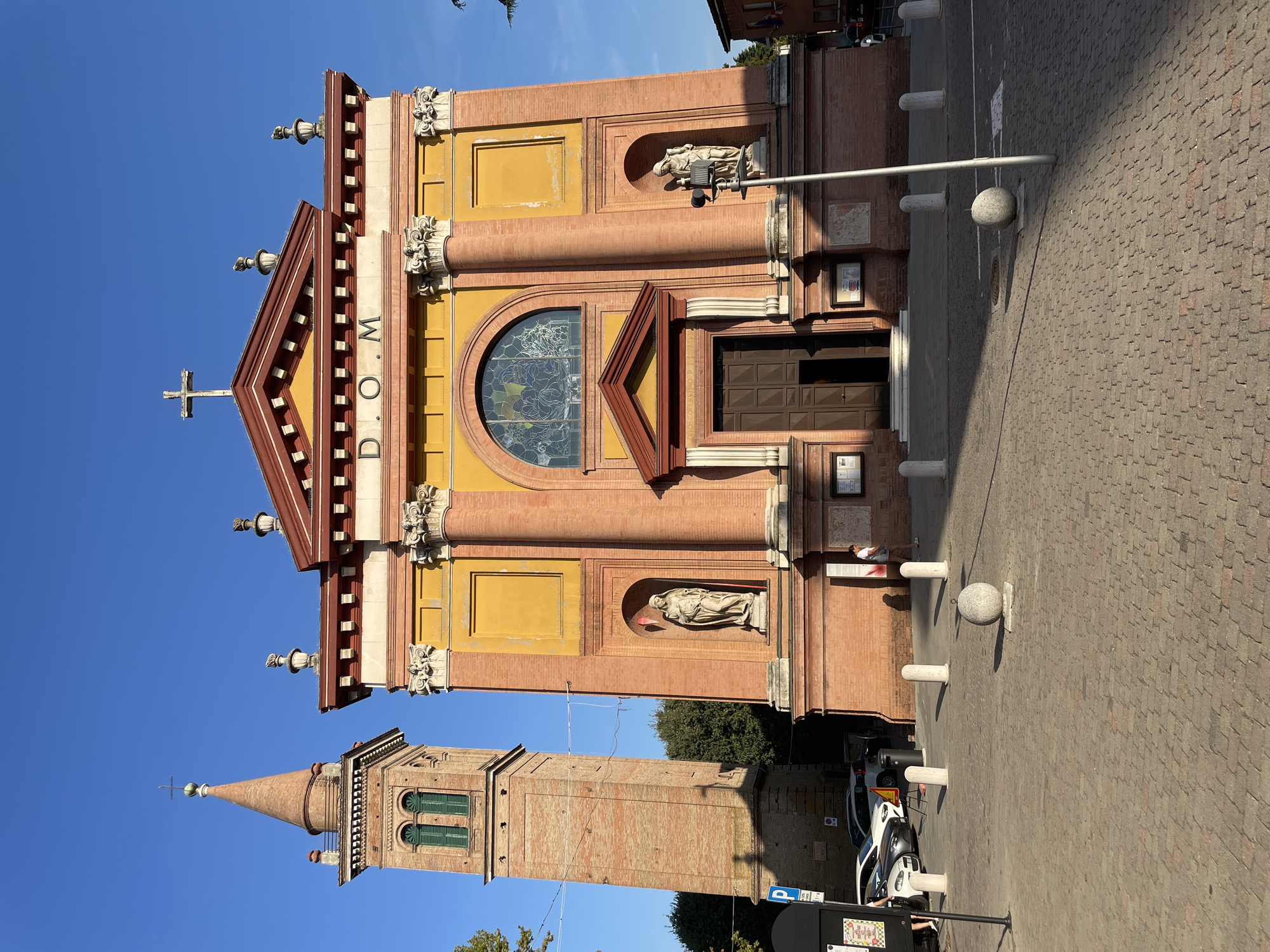 The image shows the front facade of a red brick church with a yellow painted upper section, located in Castelnuovo Rangone, Italy. A tall, slender bell tower made of red brick with green shutters stands to the left of the church. The church has a prominent entrance with a large wooden door and a stained-glass window above it. Niches on either side of the entrance contain statues. The facade is decorated with columns and pediments. The ground in front of the church is paved with cobblestones and has several white bollards and a round stone planter. A street with parked cars and a pedestrian walkway is visible to the left. The sky is clear and blue.