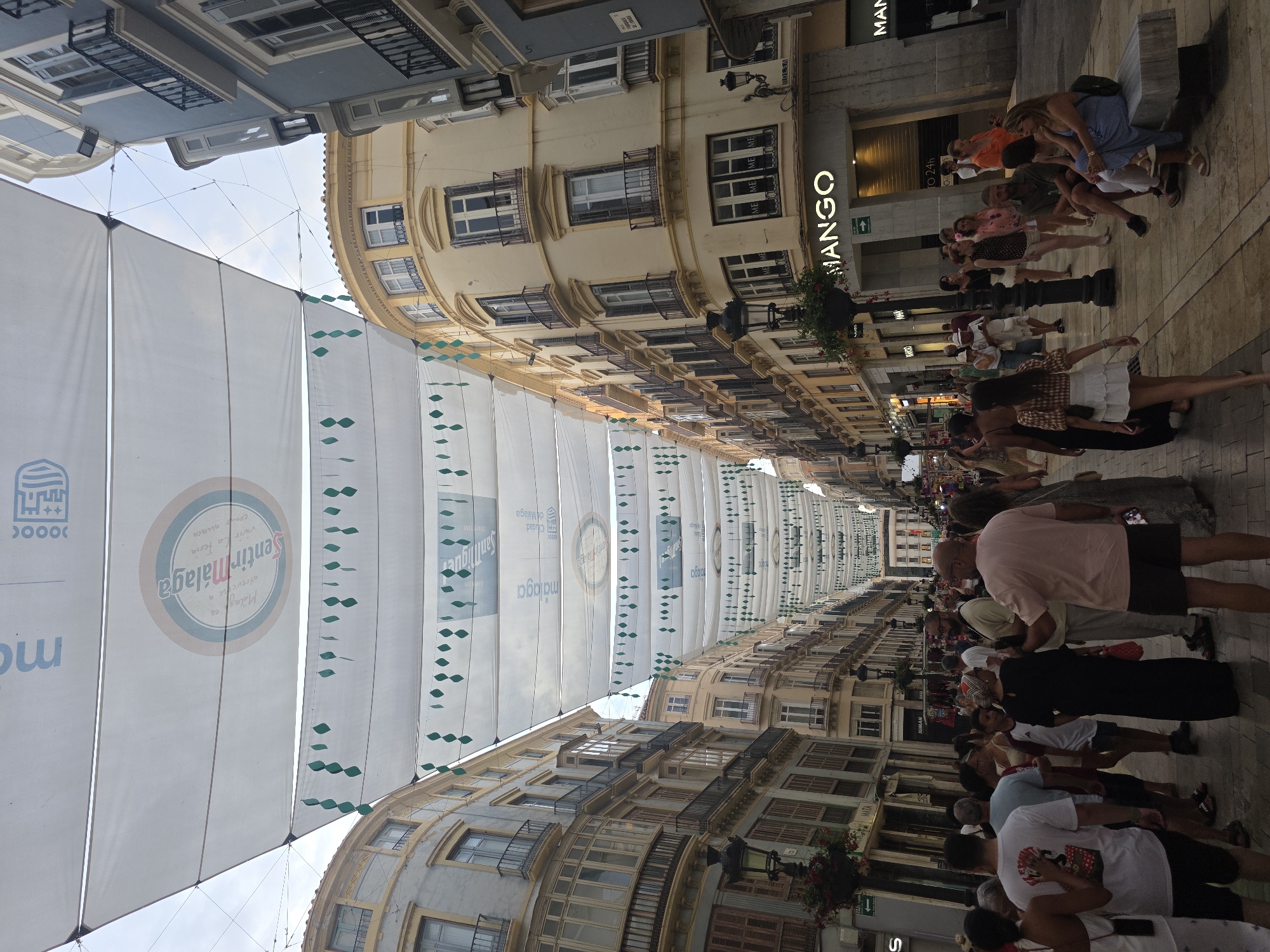 The image captures a street scene in Málaga, Spain, likely during a festival or event, given the overhead decorations. A wide street is lined with multi-story buildings on either side, showcasing a mix of architectural styles. The buildings feature balconies with ornate railings, shuttered windows, and a variety of pastel and neutral facades, including yellow, blue, and grey.
Suspended above the street are large white fabric canopies, decorated with strings of green diamond-shaped elements and various logos and text. One prominent circular logo features text that appears to include "Sentir Málaga."
The street is populated by a crowd of people of varying ages, dressed in casual summer attire. Individuals are walking, standing, and some appear to be socializing. In the foreground, a group of people are visible, including a man in a pink t-shirt and shorts, a woman in a long black dress, and others in t-shirts and shorts. Further down the street, more individuals are dispersed along the pedestrian walkway.
On the right side of the street, a store with a prominent "MANGO" sign is visible. Other shopfronts and lampposts are interspersed along the street. The ground surface appears to be paved with cobblestones or similar material. The lighting suggests it is daytime, possibly late afternoon or early evening, with soft light illuminating the scene.