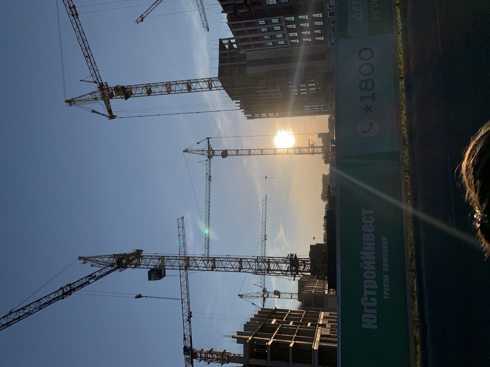The image depicts a construction site in Stavropol, Russia, under a clear blue sky at sunset. Several large yellow construction cranes are silhouetted against the sky, some with their booms extended. In the foreground, unfinished concrete buildings rise, showing the skeletal framework of apartments or offices. The setting sun casts a warm glow, with lens flares visible. A green construction fence runs along the bottom of the image, bearing white text that appears to be a company name, "ЮгСтройИнвест," and a phone number, "*1800." The fence also has other text in Russian that is partially obscured. The overall scene suggests an active construction project underway in the city.