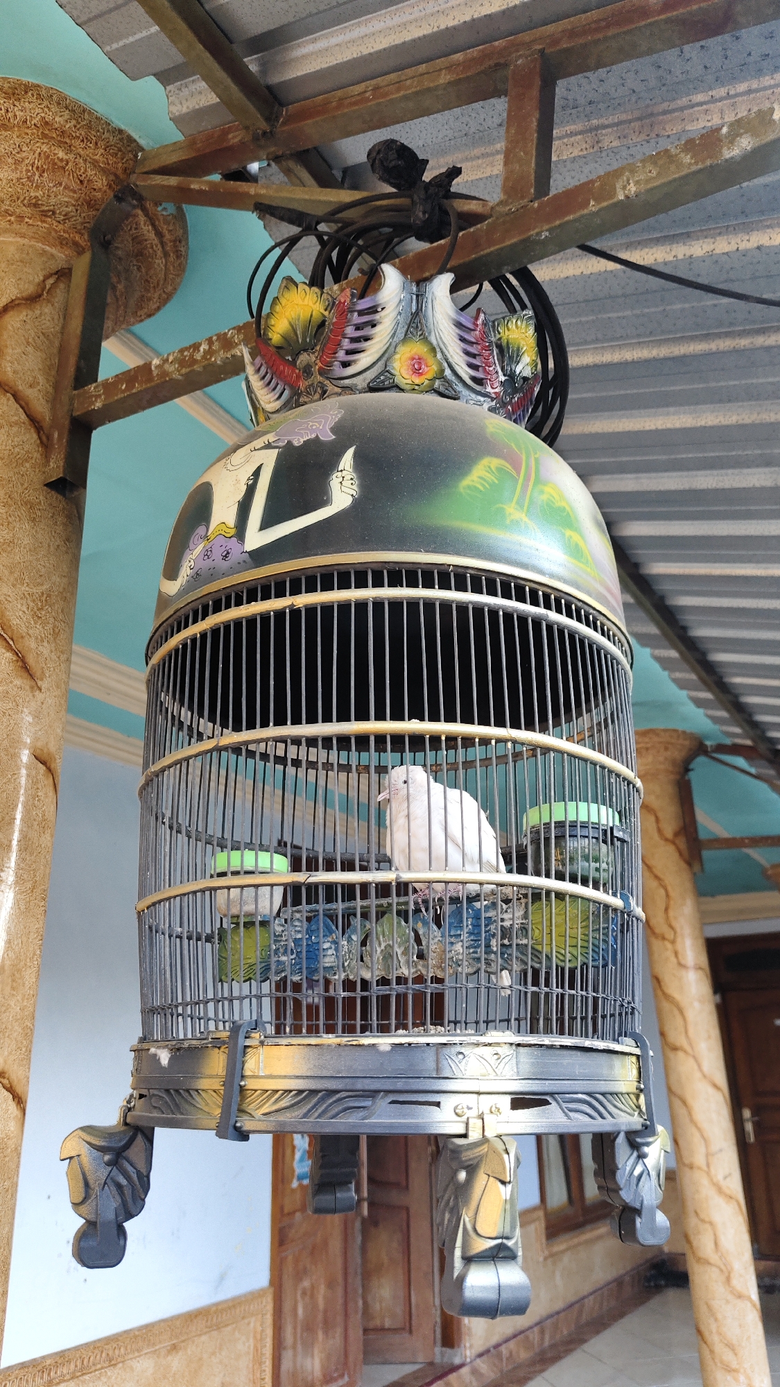 A single, white bird is perched inside a decorative birdcage hanging from a metal structure under a corrugated metal roof. The birdcage is ornate, with painted designs on its dome and carved feet. The background shows what appears to be an outdoor setting with pillars and a doorway. The scene is likely located in Galis, Indonesia, given the architectural style and possibly the type of birdcage.
