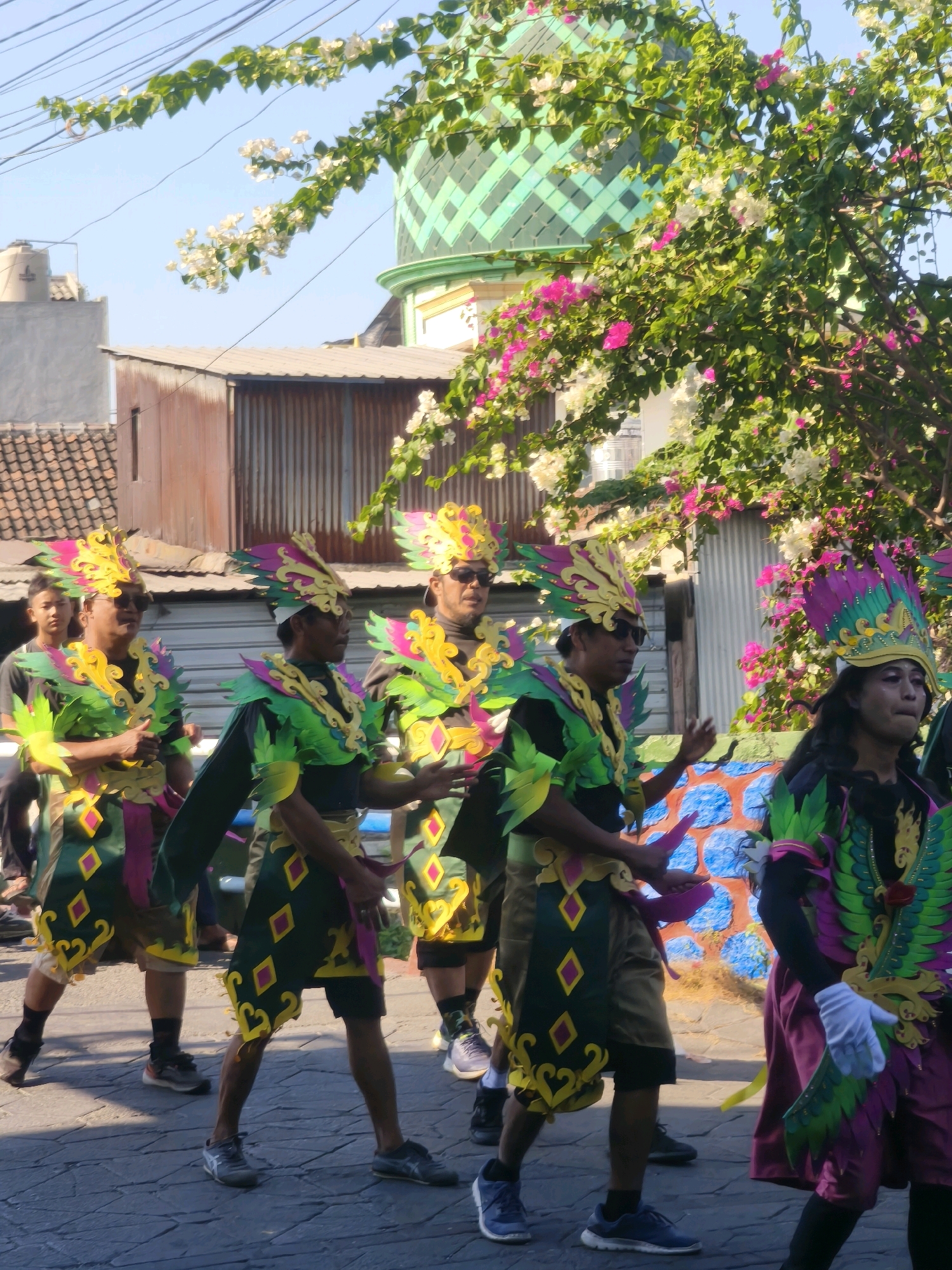 In the city of Kepuh Kiriman, Indonesia, a group of individuals dressed in vibrant, ornate costumes participate in a parade or festival. The costumes feature elaborate headdresses adorned with colorful plumes in shades of yellow, pink, and green, as well as shoulder pieces and tunics decorated with diamond patterns and fringed edges. The attire appears to be of a cultural or ceremonial nature.
The procession is taking place outdoors on a paved street, with buildings and foliage forming the backdrop. In the background, a green domed structure, possibly a mosque, is visible, partially obscured by lush green trees with abundant pink and white flowers. Electrical wires crisscross the sky above. The people in the foreground are walking forward, some with hands outstretched as if performing or interacting with an audience. Their faces are partially visible, with some wearing sunglasses. The overall atmosphere suggests a lively cultural event.