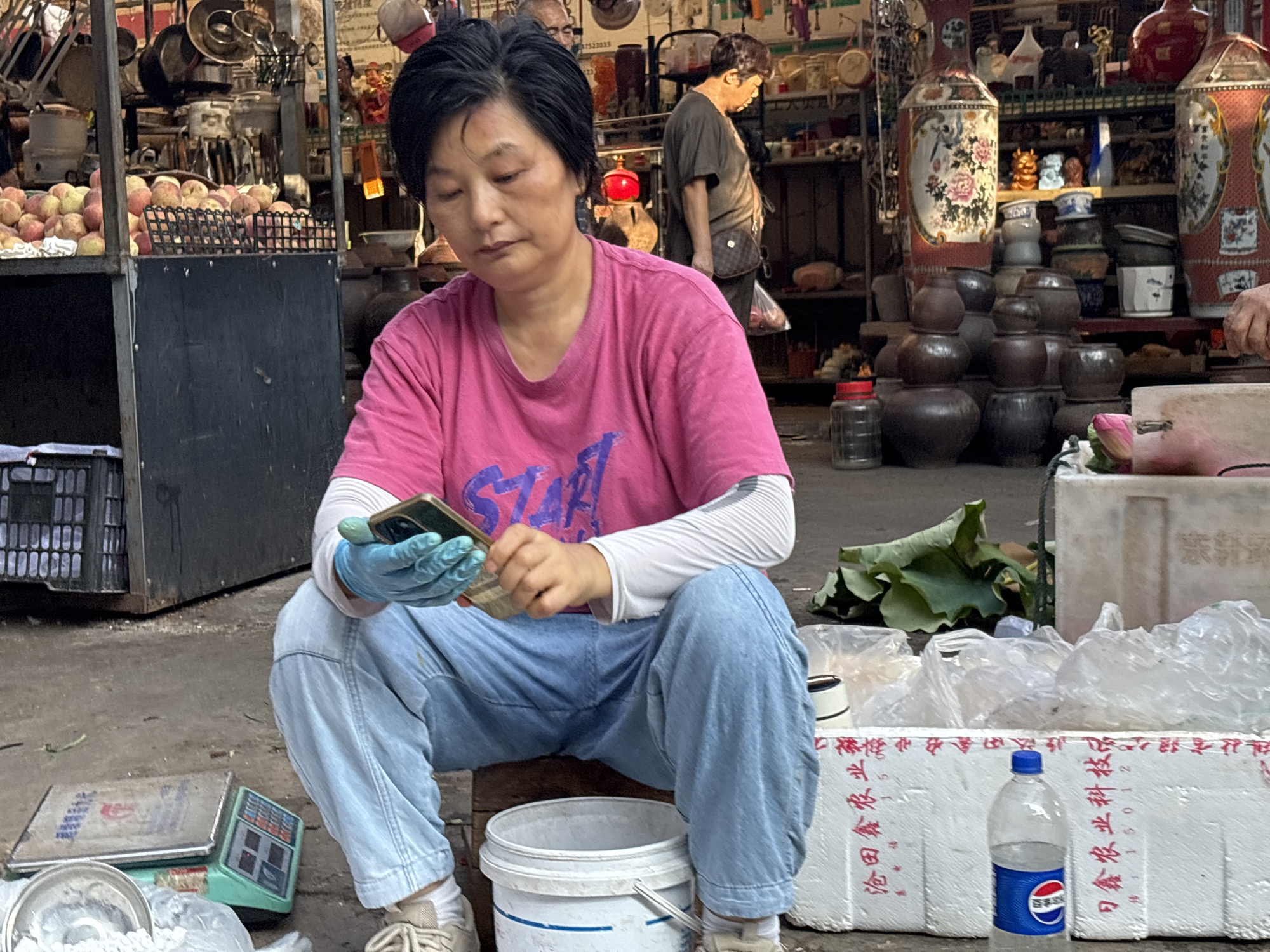 The image depicts an outdoor market scene in Jiangjin, China. In the foreground, a woman wearing a pink t-shirt and blue jeans, with blue gloves on her hands, is seated on the ground. She is looking at her mobile phone, holding it with both hands. Beside her, on the ground, are various items including a scale, a white bucket, and plastic-wrapped goods. In the background, stalls are filled with produce, pottery, and other merchandise. Another person is visible in the background, partially obscured. The overall environment suggests a busy marketplace with an array of goods and vendors.