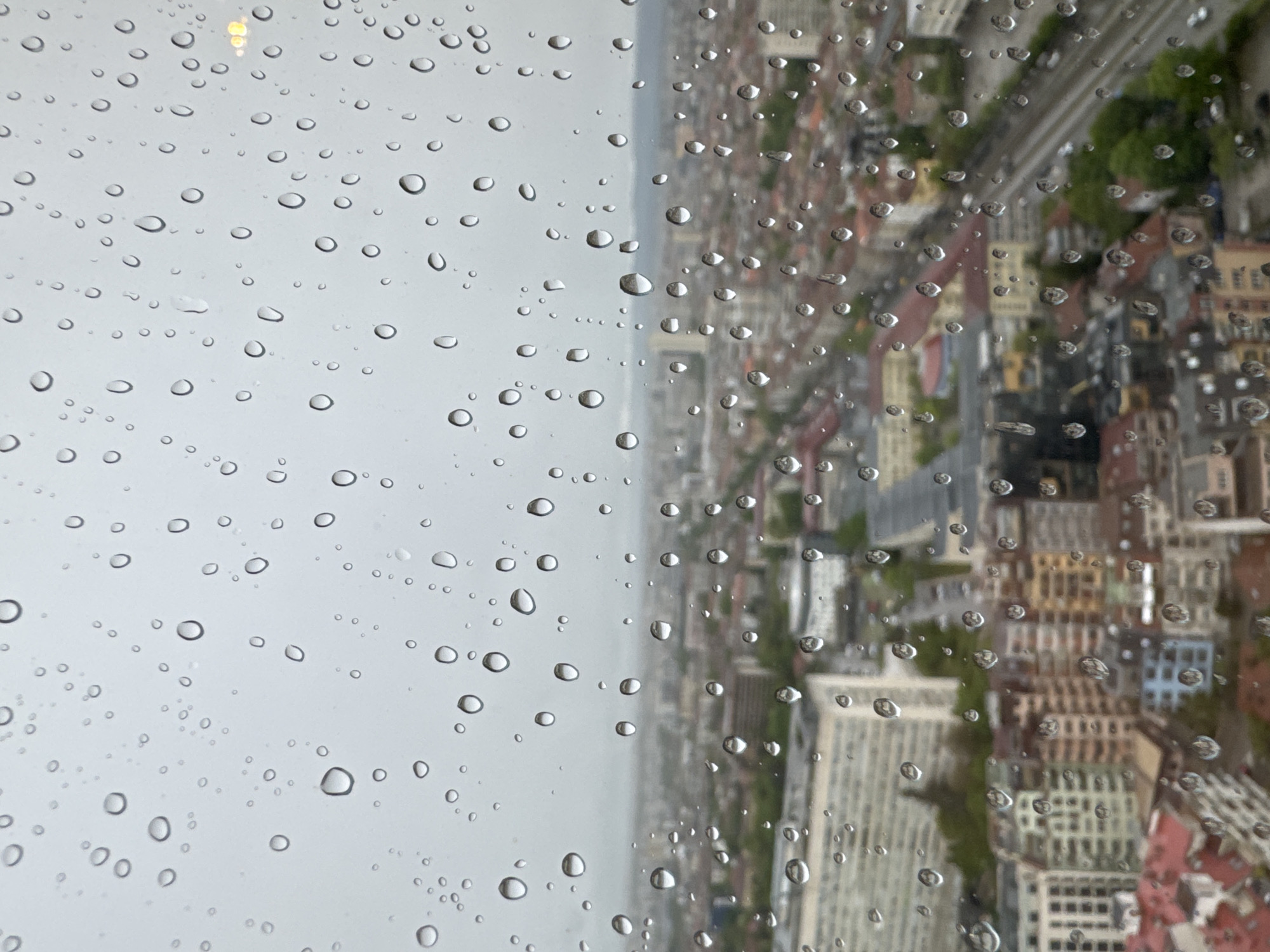 The image displays a high-angle view of Sofia, Bulgaria, seen through a window covered in numerous raindrops. The scene is bisected by an indistinct vertical line, suggesting a window frame or the edge of a clear view.
On the left side of the frame, large and small water droplets cling to the glass, set against a soft, light gray background, indicative of an overcast sky or dense fog. The individual droplets vary in size, some perfectly spherical, others slightly elongated, each reflecting and refracting the ambient light, creating tiny, bright highlights and darker outlines.
The right side of the image reveals a densely packed urban landscape, heavily blurred and distorted by the wet glass and the distance. Colorful buildings with varied rooflines (including reds, browns, oranges, and grays) and facades are visible through the shimmering curtain of water. Patches of green, likely trees or parks, are interspersed among the structures. An elevated grey structure, possibly a highway or rail line, stretches across the mid-ground.
The visual effect of the raindrops on the window blurs the cityscape into an impressionistic pattern, contrasting sharply with the clear, detailed forms of the water droplets themselves. The weather is clearly rainy or has just rained, and the diffused lighting suggests a cloudy daytime. No visible text is present in the image.