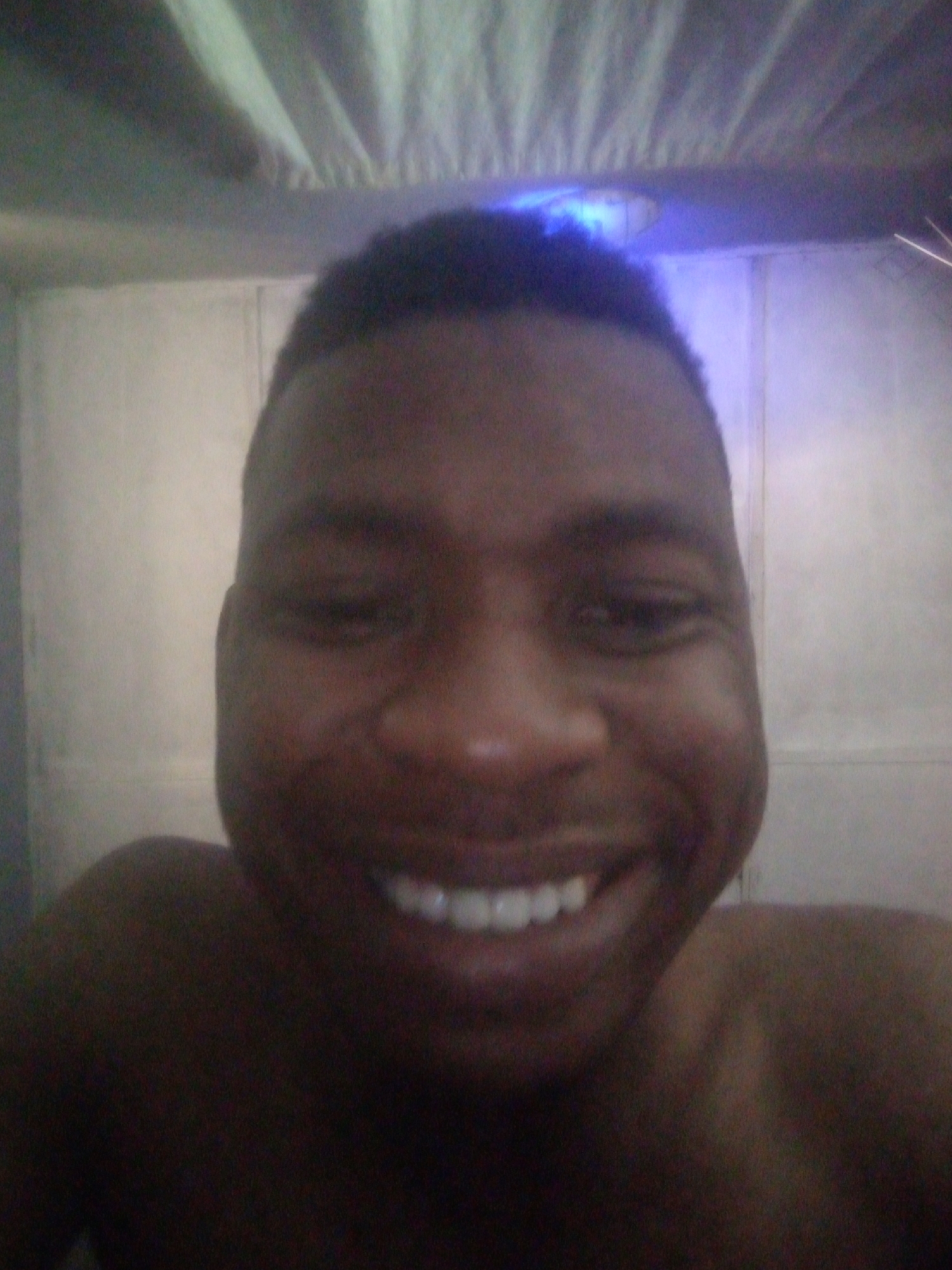 A medium-close selfie captures a dark-skinned man with short, dark hair, smiling broadly. The background is an off-white wall illuminated by blue light, suggesting an indoor setting. The man is shirtless. The location, Ikorodu, Nigeria, is not directly indicated in the image and must be deduced from external information.