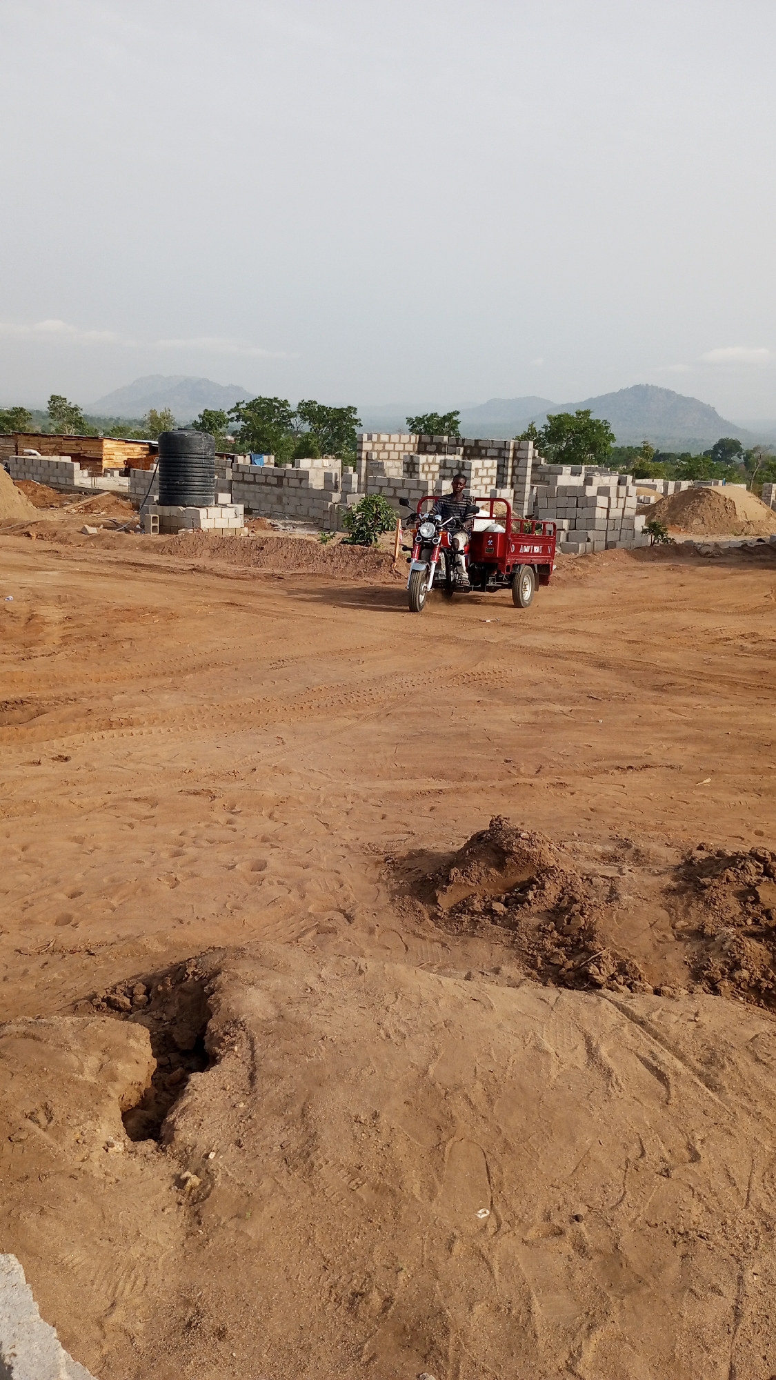 The image depicts a construction site in Zango-ndaji, Nigeria. A man is riding a red and white three-wheeled motorbike with a small trailer attached. The motorbike is heading towards the viewer. The ground is mostly dirt, with piles of sand and mud. In the background, there are unfinished structures made of grey cinder blocks. A black water tank and stacks of lumber are also visible. The background includes green trees and hills under a cloudy sky. The overall scene is dusty.