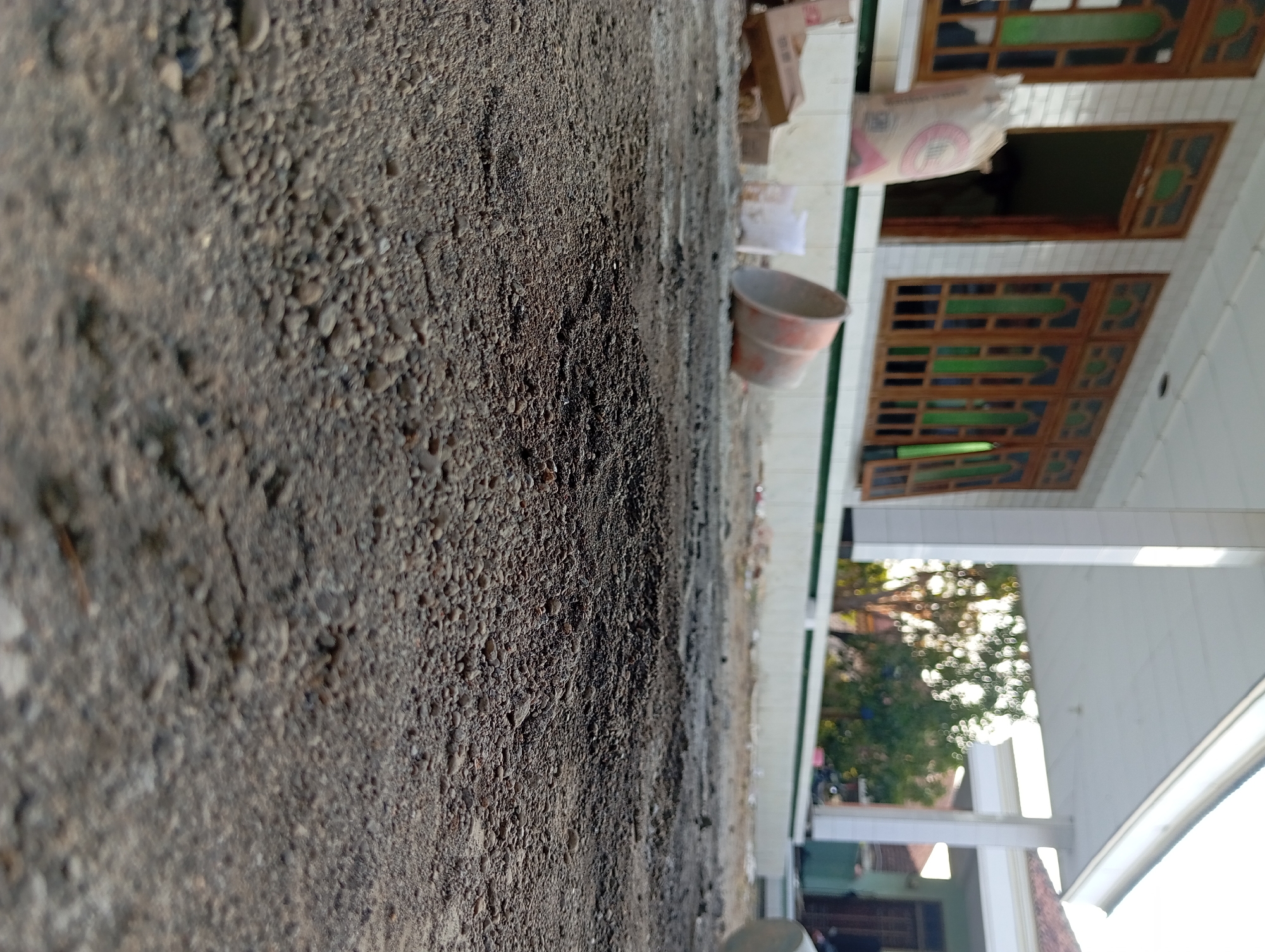 The image depicts an outdoor construction or renovation scene, likely in front of a building with white tiled walls and wooden-framed windows. The foreground is dominated by loose gravel and soil, suggesting the ground is being prepared. In the mid-ground, a large sack, possibly containing building materials like cement, is visible leaning against the building. There are also some cardboard boxes and a white plastic container. A large, orange bucket or tub lies on its side. The background shows more of the building's exterior, including pillars supporting an overhang. Trees and foliage are visible further in the background, along with other structures. The overall impression is one of ongoing work, with materials and debris scattered around a partially finished or developing area. The location is specified as Padurungan, Indonesia.