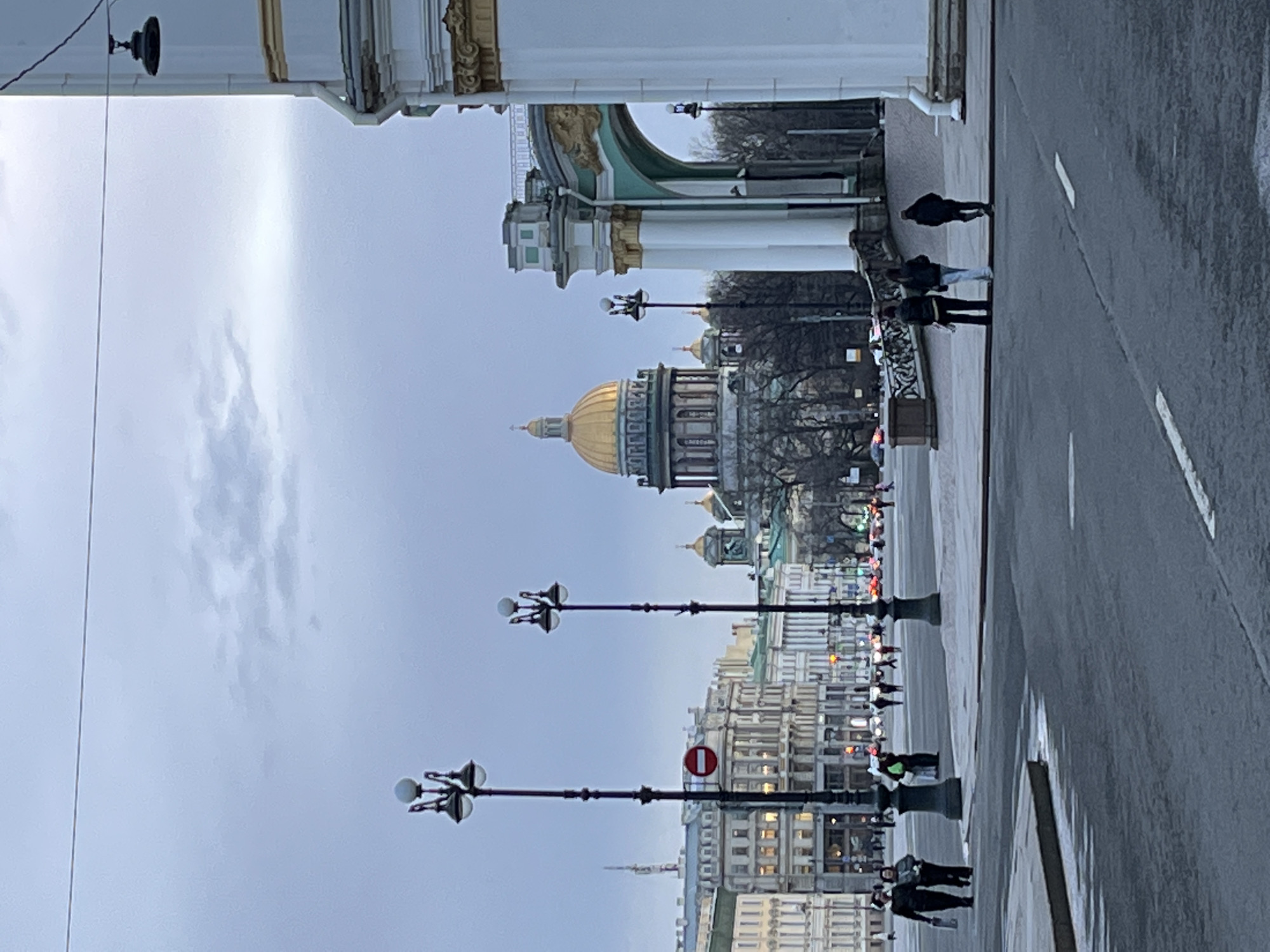 The image shows a street scene in Saint Petersburg, Russia. In the background, a large golden dome dominates the skyline, likely belonging to Saint Isaac's Cathedral. The foreground features a wet road with white lane markings, and a sidewalk where several pedestrians are walking. Along the sidewalk, there are ornate street lamps. Above, architectural details of a building or archway are visible. The sky is overcast, suggesting a rainy or cloudy day.