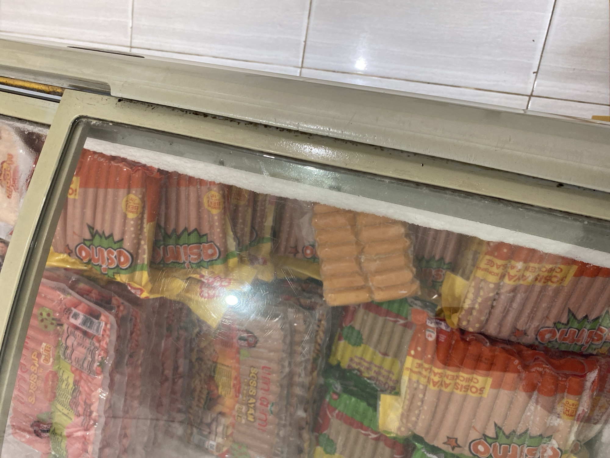 The image shows a close-up view of a commercial freezer chest containing several packages of sausages. The freezer is a light beige color and has a glass lid, through which the products are visible. The sausages are packaged in clear plastic with various colorful labels. Some labels are red and yellow with the brand name "Sosis Ayam" (Chicken Sausage) and other text in Indonesian. There are also packages with green and yellow labels featuring a stylized green star and the word "Osino." The sausages themselves appear to be thin and reddish-pink in color. Some packages are stacked on top of each other, and the freezer appears to be full. The interior of the freezer is frosted in places, indicating its low temperature. The lighting inside the freezer is bright, possibly from overhead lights. The background shows a tiled floor, likely in a commercial setting. The overall scene suggests a retail or food service environment where frozen food products are stored. The location in Payatieng, Indonesia, is context provided externally and not directly discernible from the image itself.