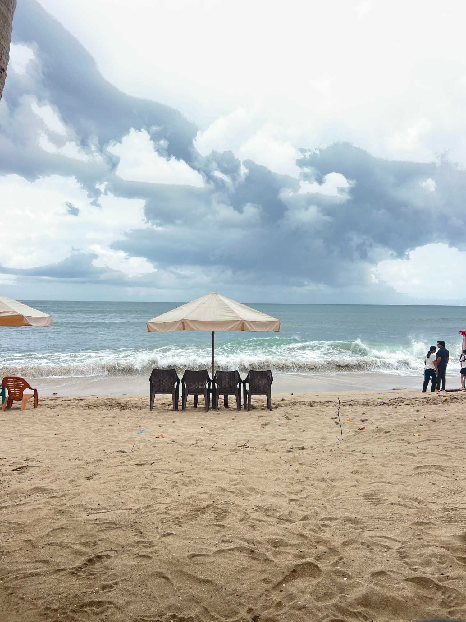 This image depicts a beach scene in Kuta, Bali, Indonesia, under a cloudy sky. In the foreground, a sandy beach is visible, with footprints and scattered debris like small sticks and possibly plastic fragments. Several beach chairs are arranged on the sand. To the left, a light brown plastic chair stands alone. In the center, four dark plastic chairs are lined up under a light-colored umbrella. To the right of the chairs, a second light-colored umbrella is set up.
Further back, the ocean stretches out to the horizon. Waves are crashing onto the shore, creating white foam and spray. The water appears a shade of blue-green.
On the beach, near the umbrellas, a group of people are visible. Two individuals, a man and a woman, are standing close together, facing the ocean. The man is wearing a dark shirt and pants, and the woman is in a white top and dark pants with a green stripe. To their right, two other figures, possibly children, are also present. The overall atmosphere suggests a somewhat overcast day at a popular tourist beach.
